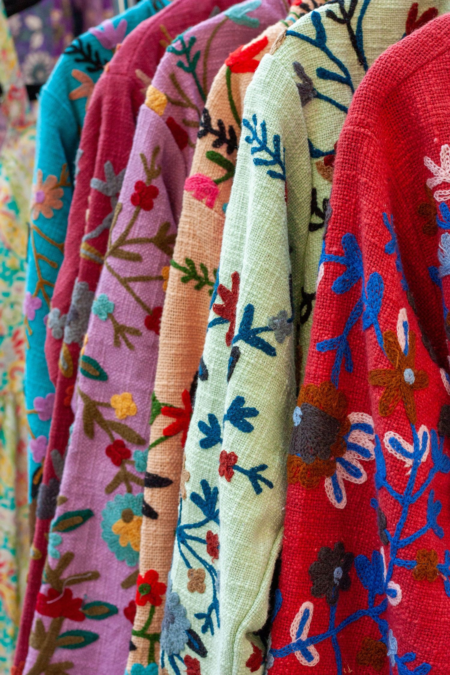 Colorful embroidered floral jackets hanging on a rack, featuring vibrant patterns in pink, red, green, and blue hues.