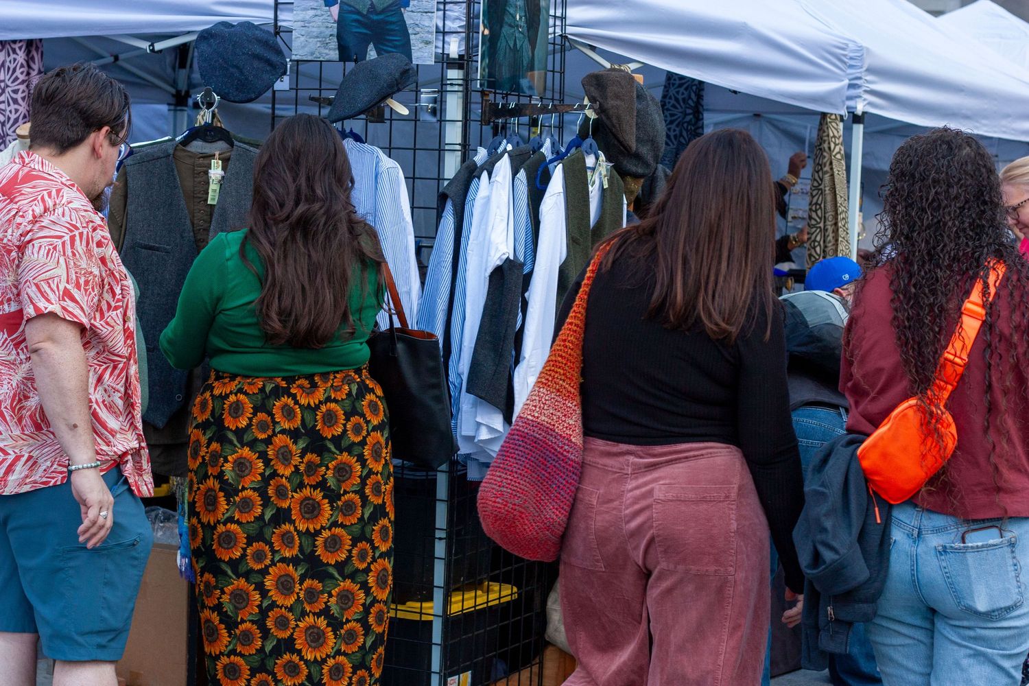 People browsing clothing racks at an outdoor market, with women in colorful outfits shopping under white canopy tents.