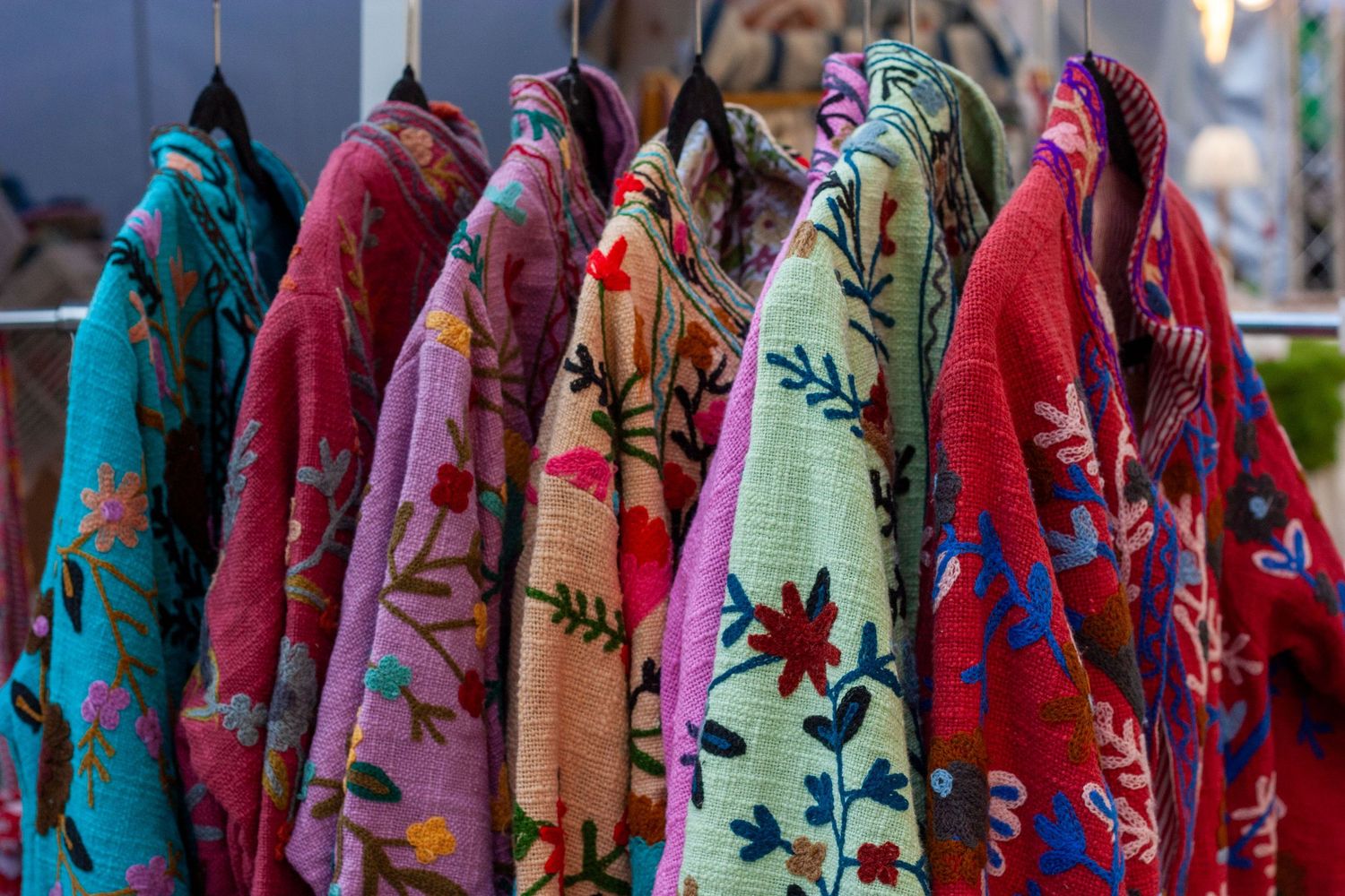 Colorful embroidered floral jackets hanging on a rack at a market, featuring vibrant patterns in pink, blue, and red.