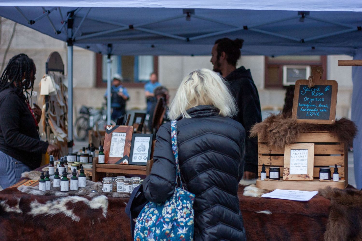 Shoppers browse handmade goods at an outdoor market stall with small bottles and rustic wooden displays under a blue tent.