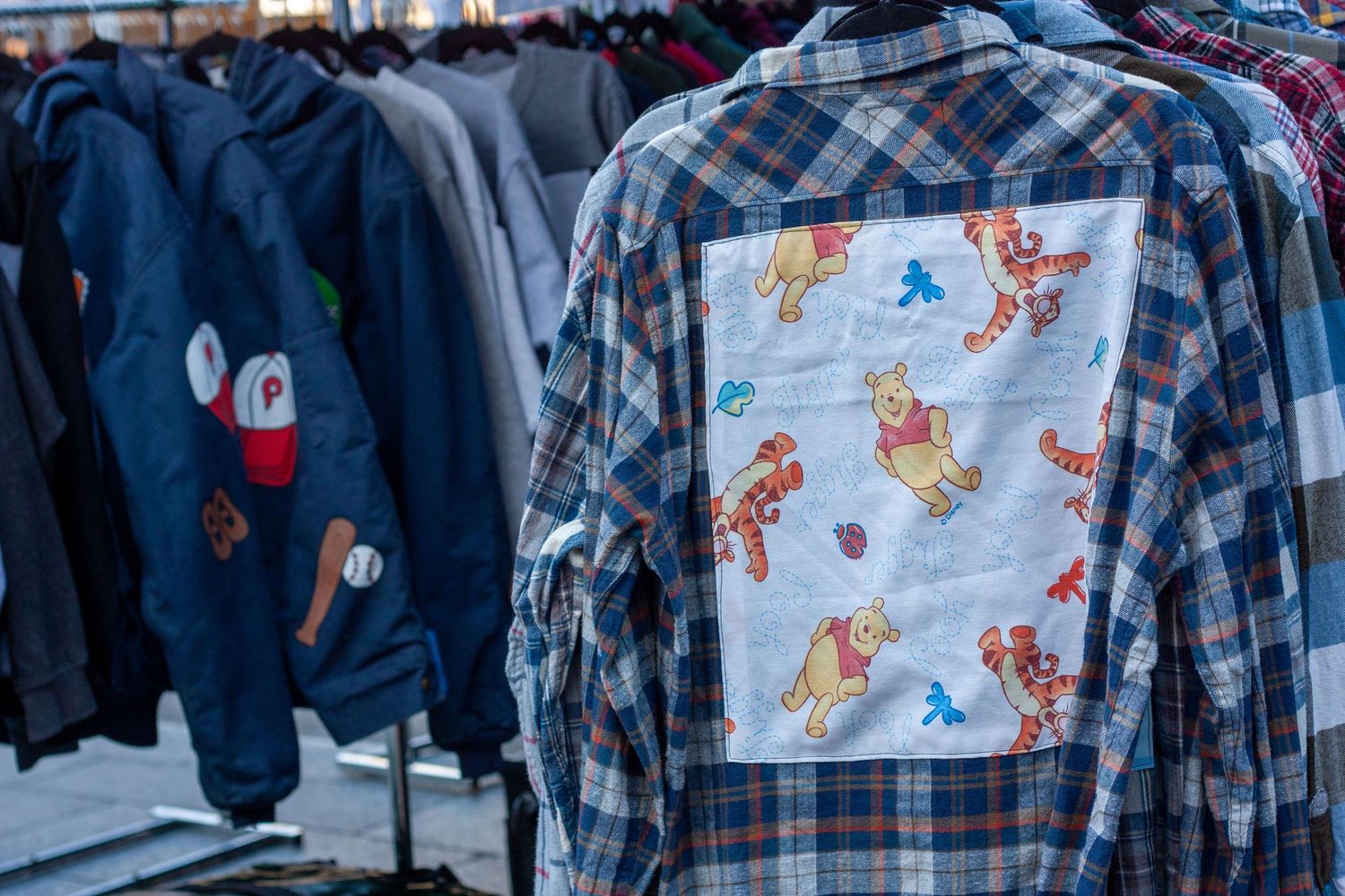 Plaid flannel shirts with Winnie the Pooh patches displayed on a clothing rack at an outdoor market.
