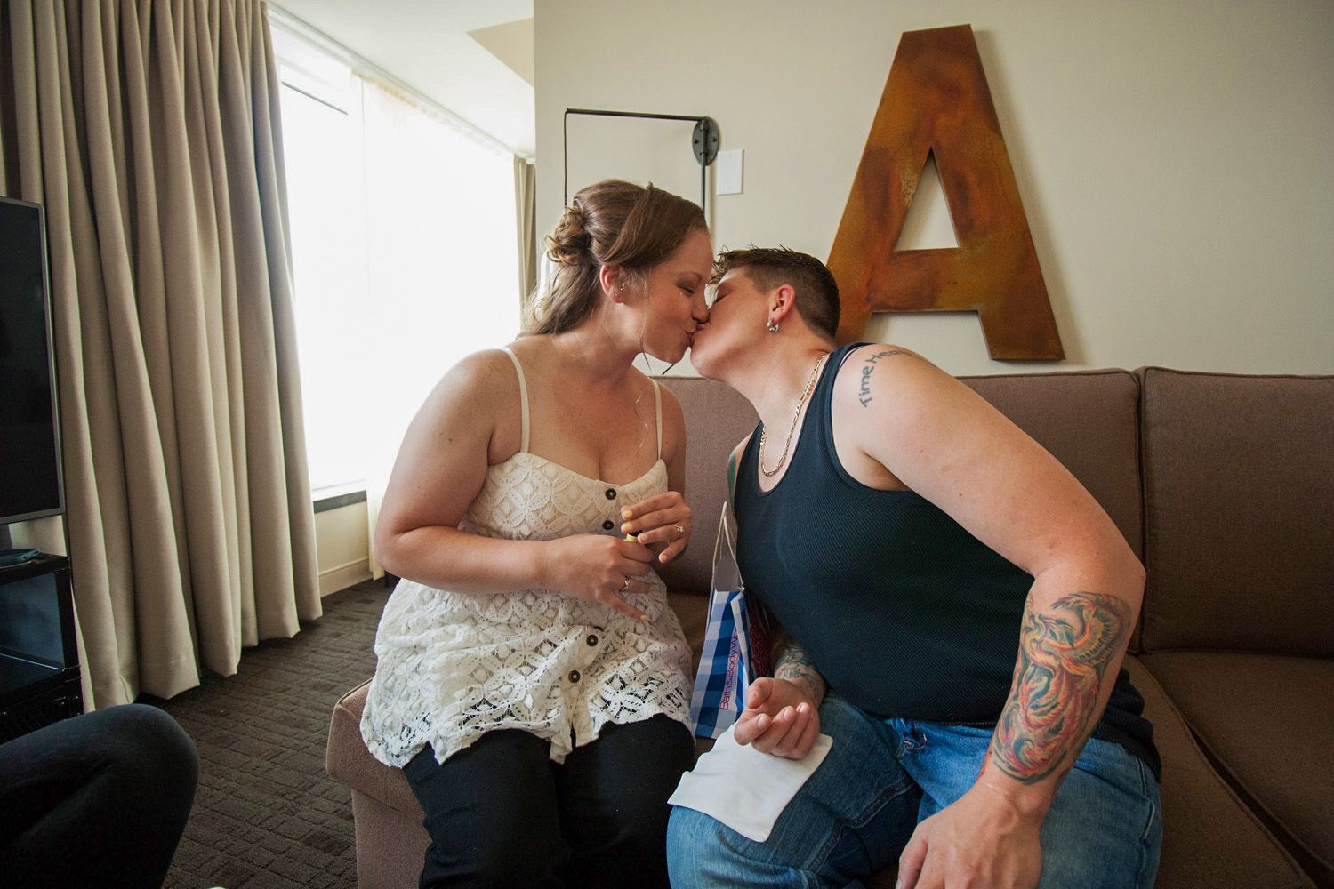 Two people sharing a kiss on a couch in a hotel room, one with tattoos, holding papers near a wooden letter A.
