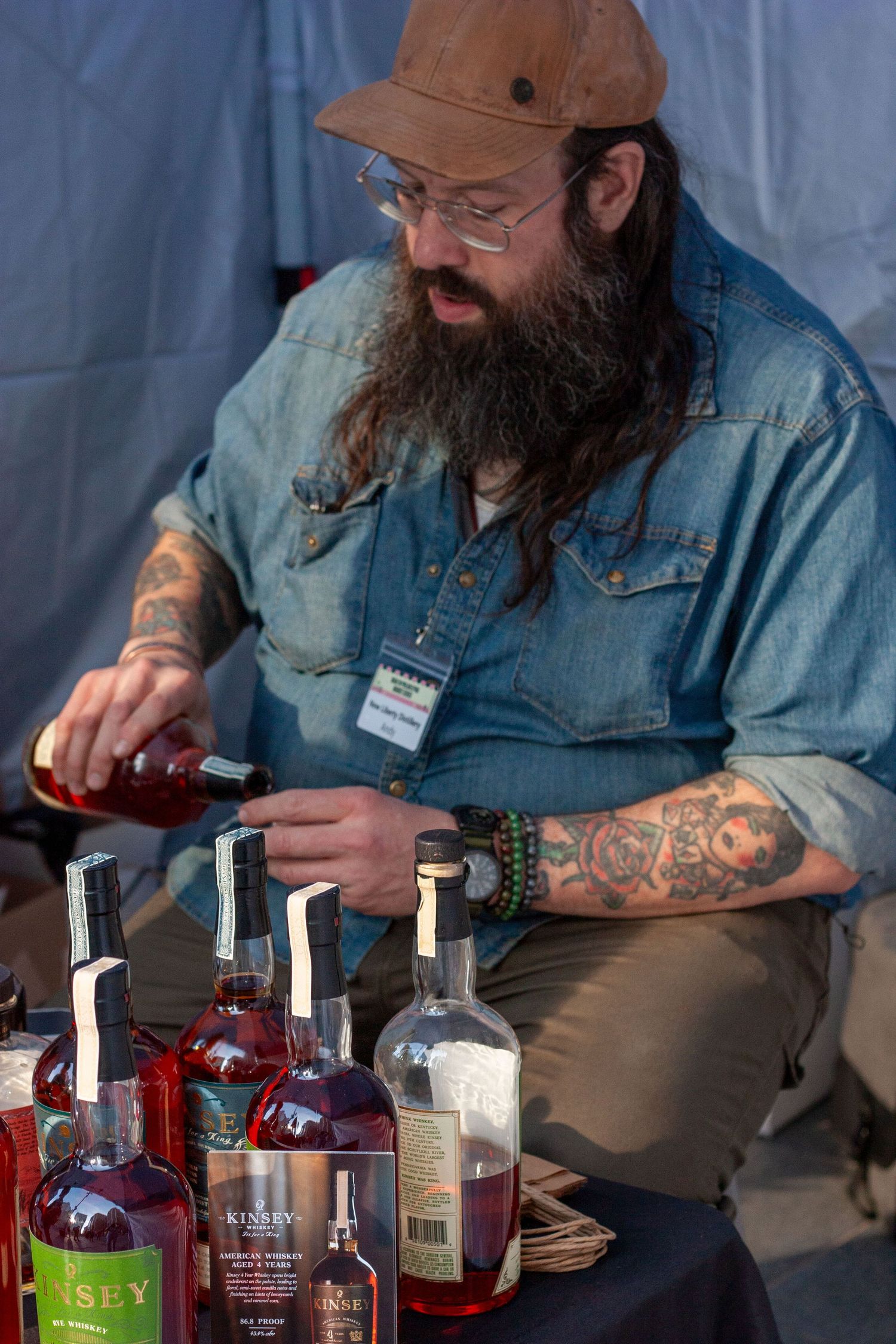 Tattooed bearded man in denim shirt and cap pouring whiskey at a bourbon tasting event with multiple bottles displayed.