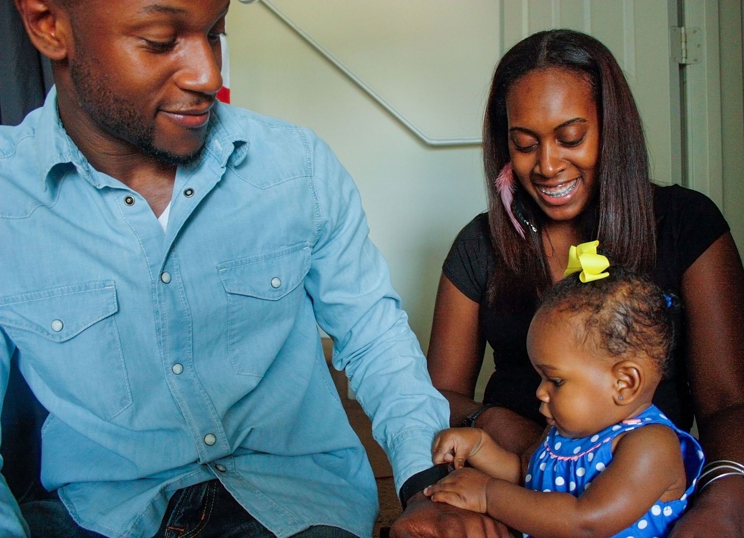 A happy Black family with parents smiling at their baby girl in a blue polka dot dress with a yellow bow.