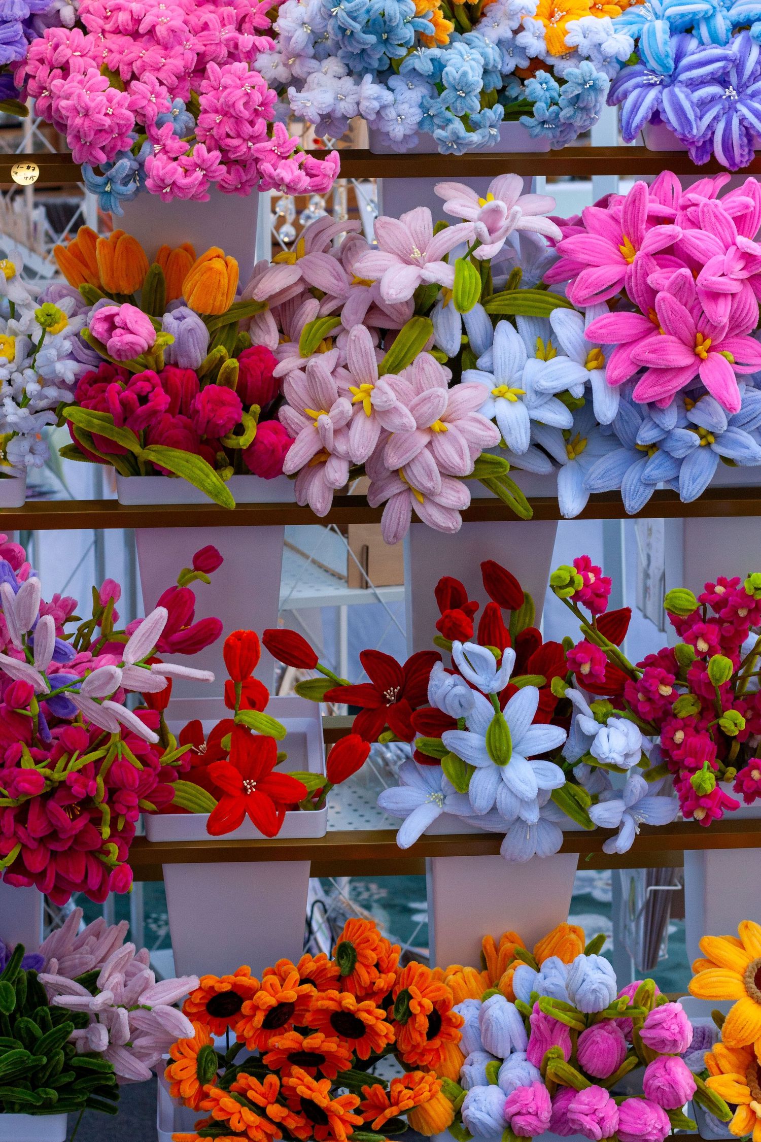 Colorful artificial fabric flowers displayed on shelves, featuring pink, blue, red, orange, and white blooms in a market.