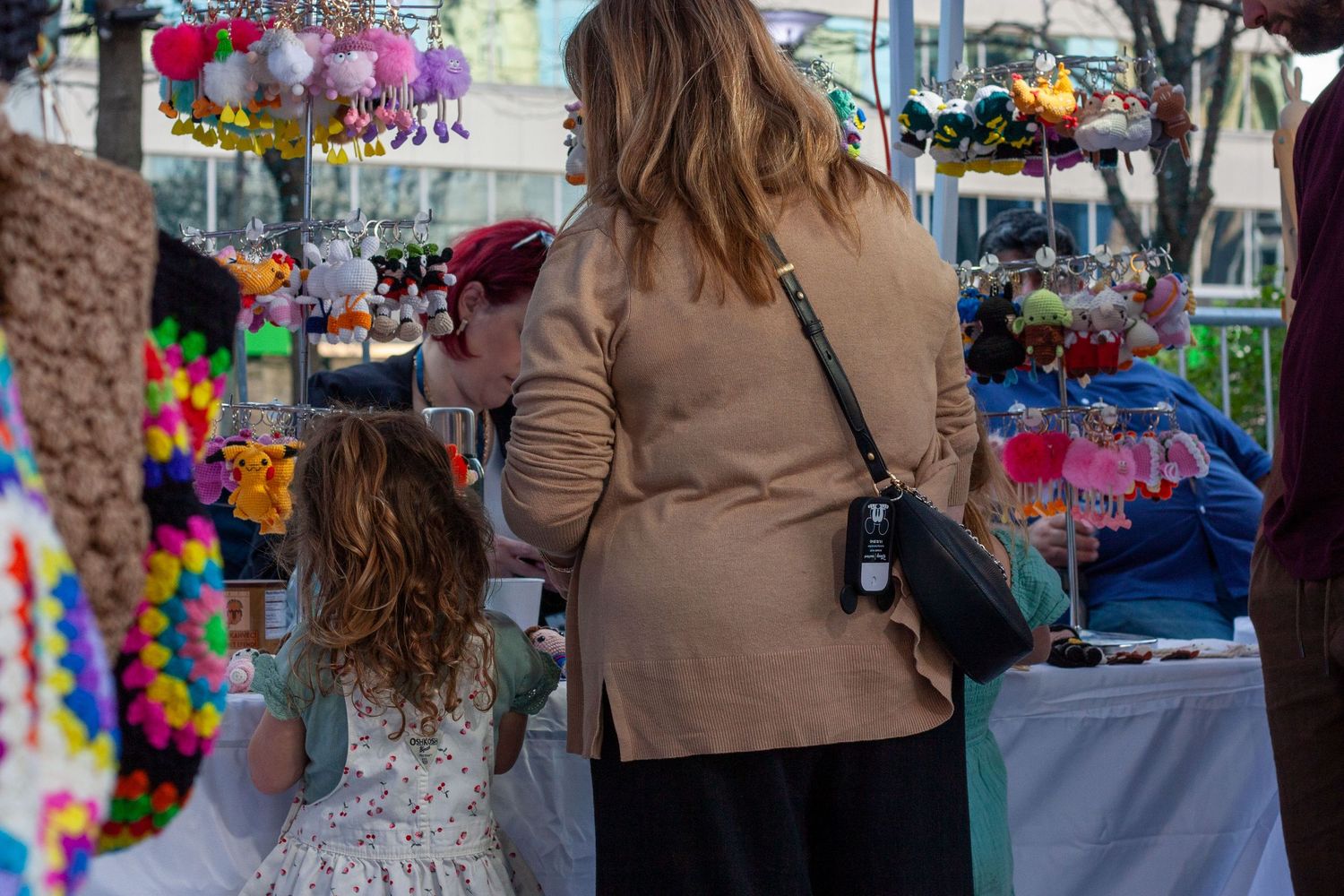 Woman and young girl shopping at colorful outdoor craft market stall with hair accessories and handmade goods.