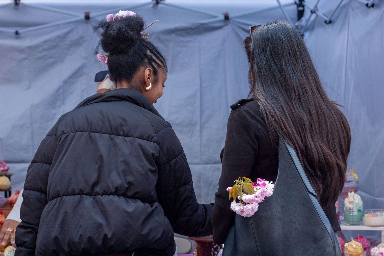 Two women holding hands and flowers stand with their backs to camera near a memorial tent.