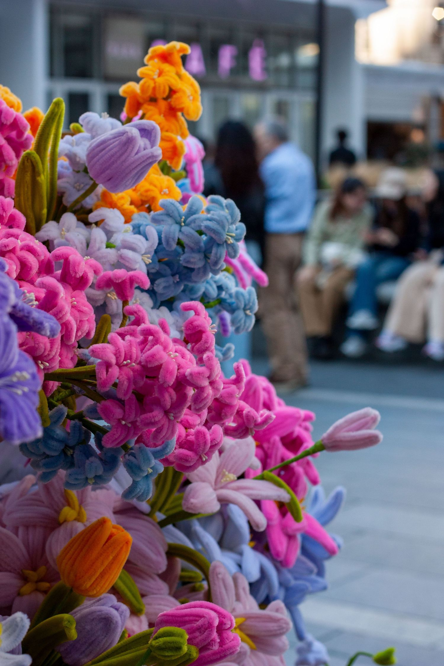Colorful handmade crochet flowers in pink, blue, purple, and orange displayed outdoors with blurred people in the background.