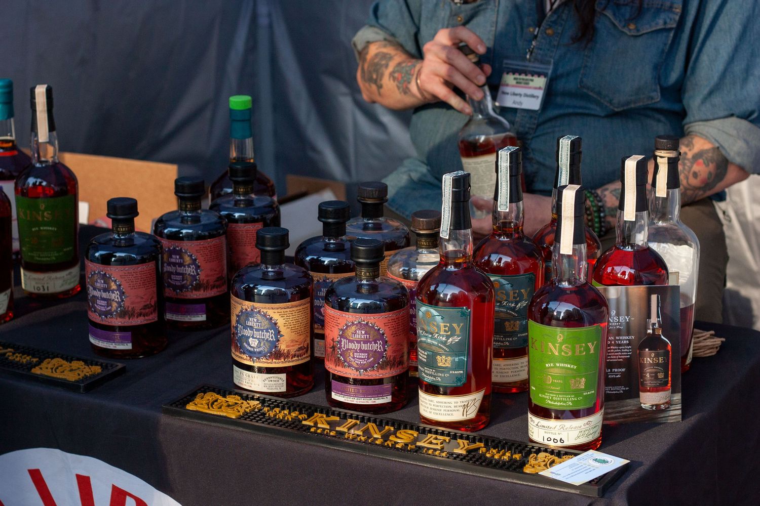 Assorted craft spirits bottles displayed on a table at a tasting event, with a tattooed person standing behind.