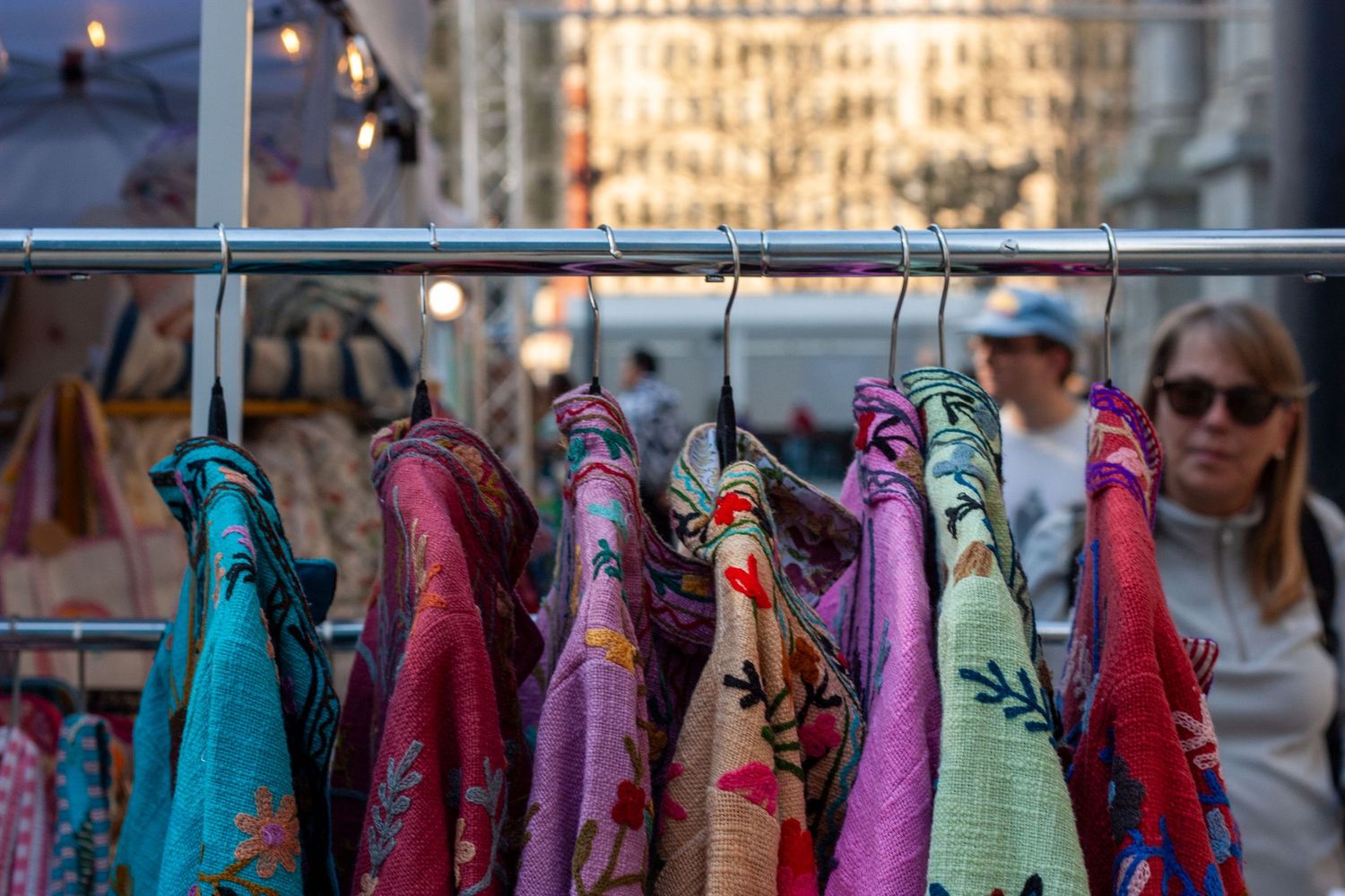 Colorful floral scarves and garments hanging on a rack at an outdoor market with shoppers in the background.