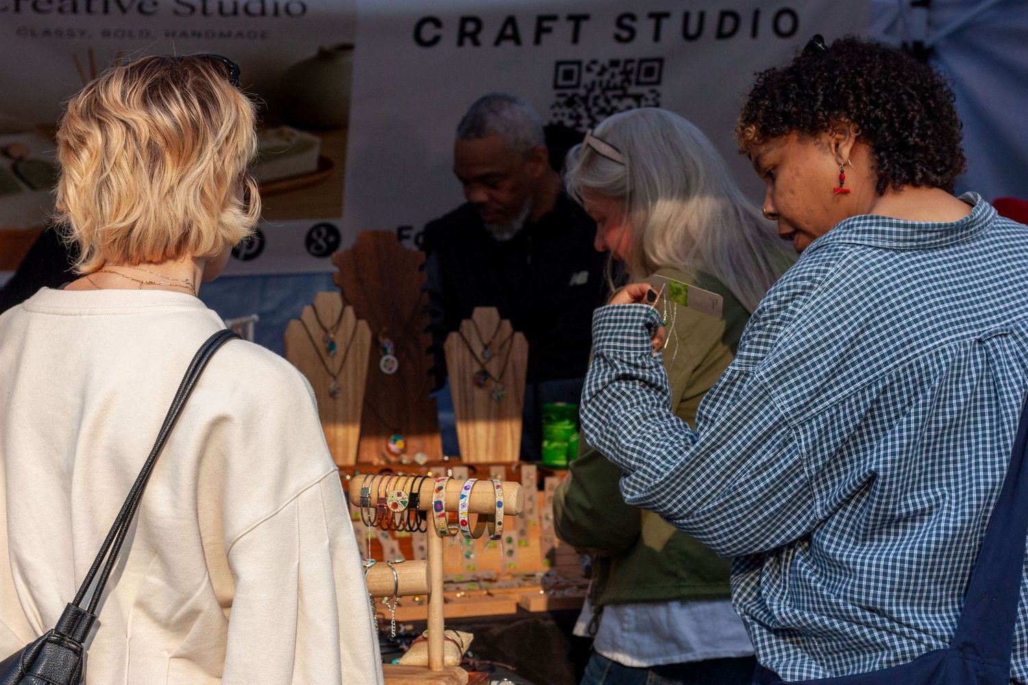 People browsing handmade jewelry at a craft studio market stall with display stands and a QR code sign.