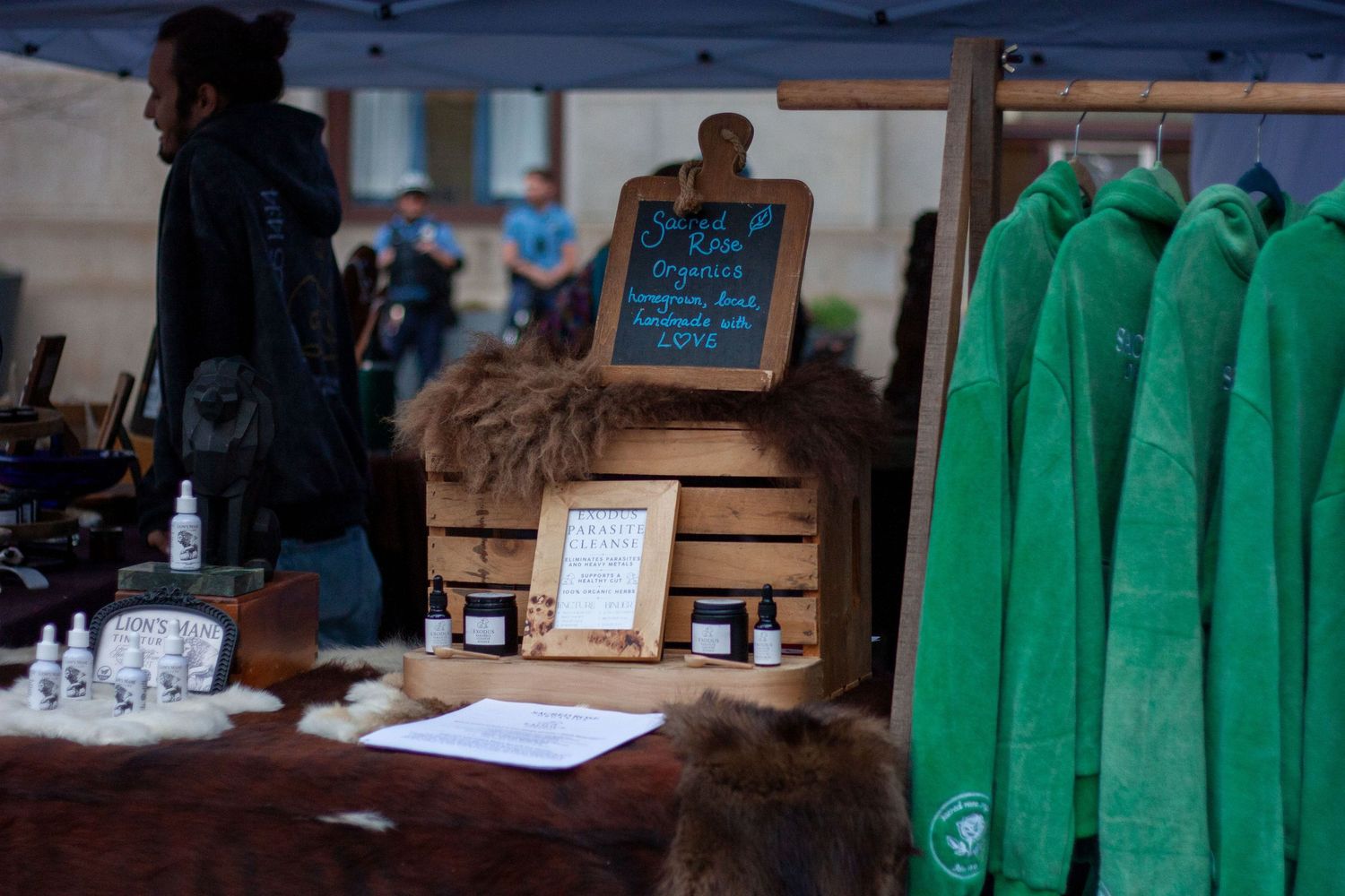 Outdoor market stall displaying organic products, wooden crates, a chalkboard sign, and green fleece jackets hanging on a rack.