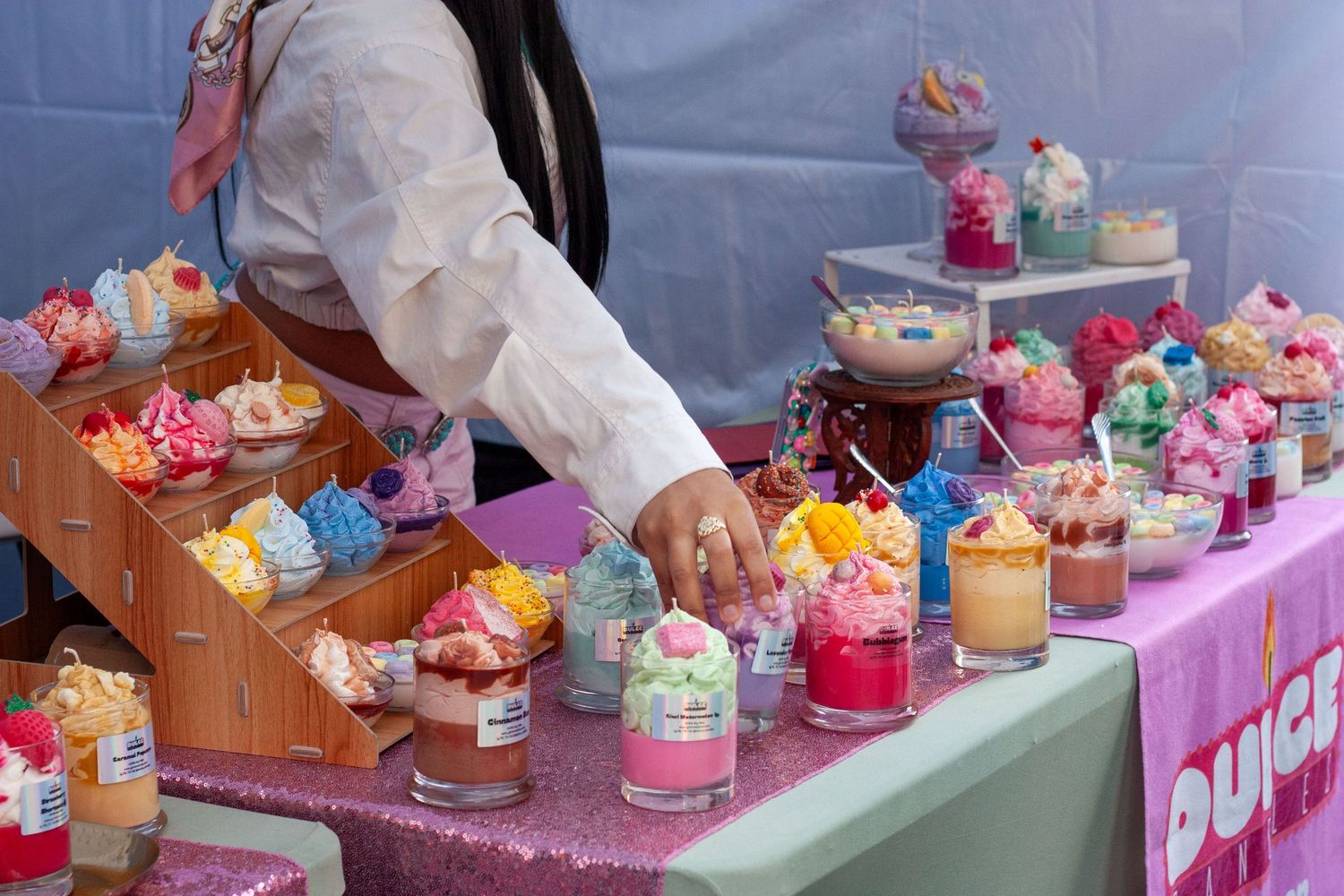 Colorful candles displayed on a glittery pink table at a craft fair, styled to resemble desserts and cupcakes.