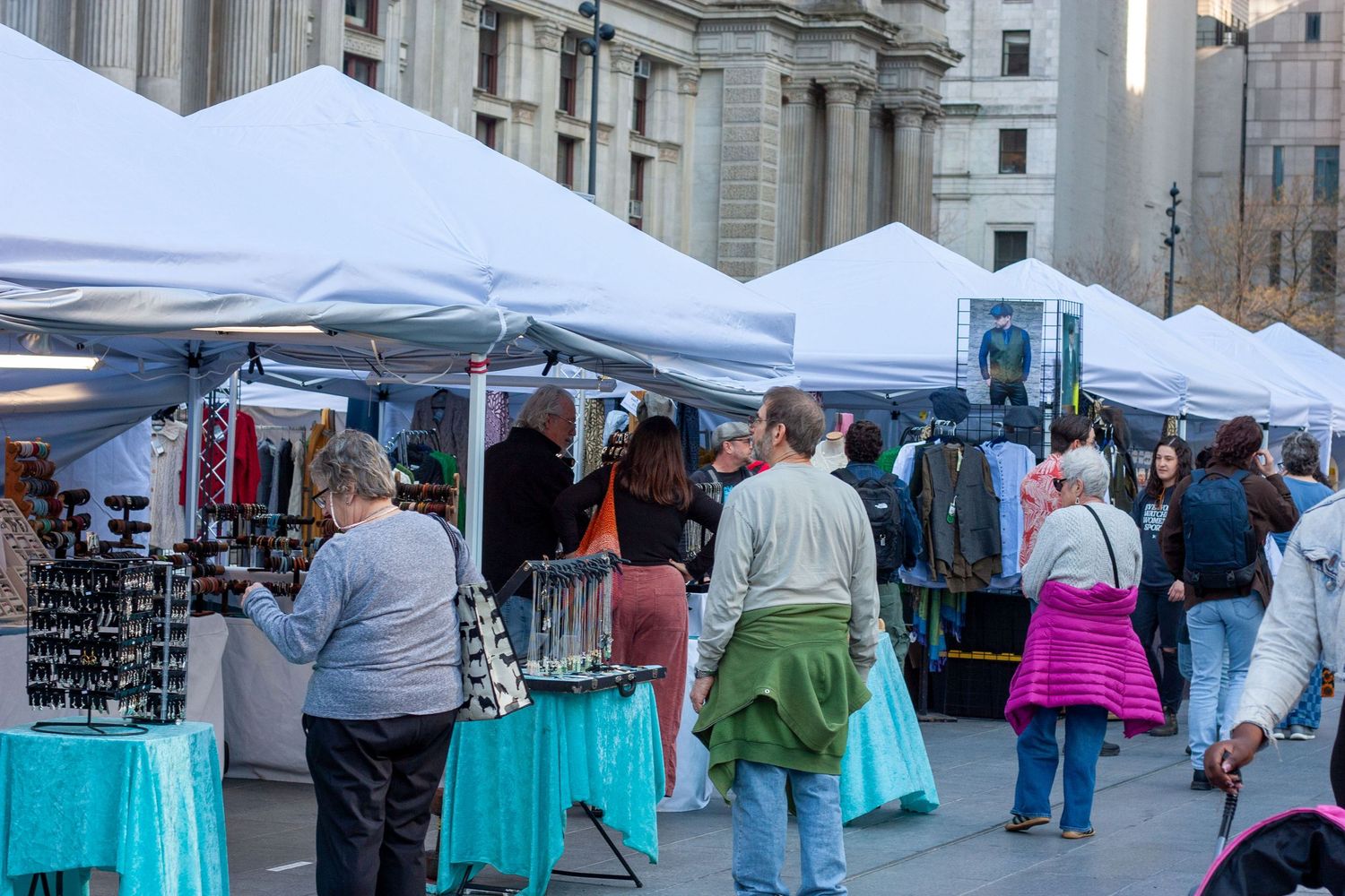 Outdoor market with white canopy tents, vendors, and shoppers browsing goods on a city plaza.