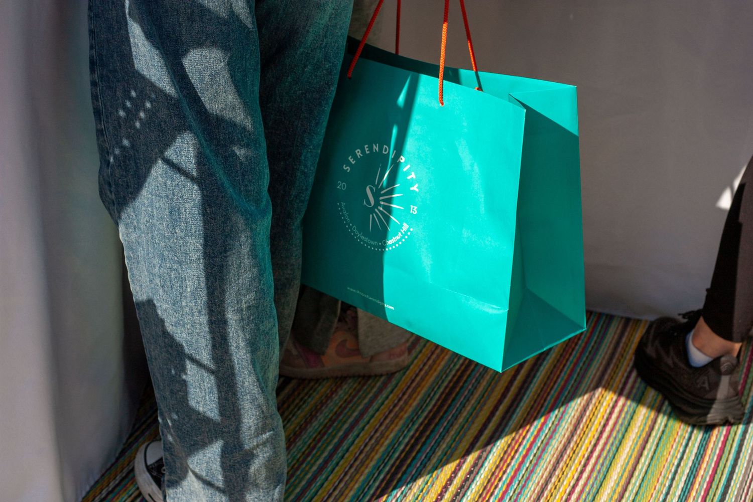 Teal luxury shopping bag with logo hanging in sunlight on colorful striped carpet floor.