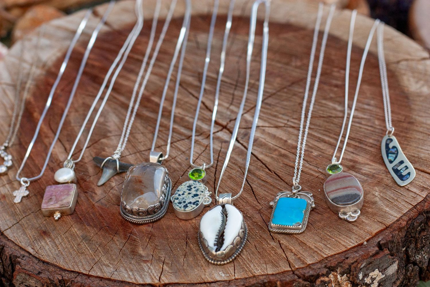 Collection of silver chain necklaces with various gemstone and shell pendants displayed on a rustic wooden stump.