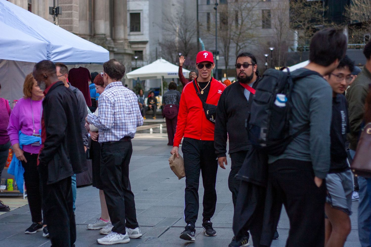 Crowd of people walking through an outdoor urban plaza with white tents and a man in a red jacket.