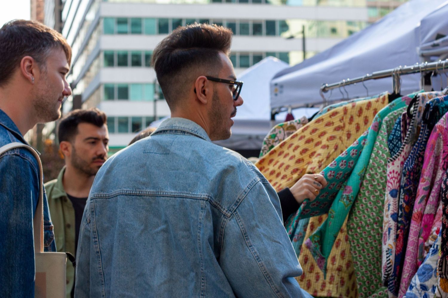 Three men browsing colorful clothing on a rack at an outdoor market in an urban setting.
