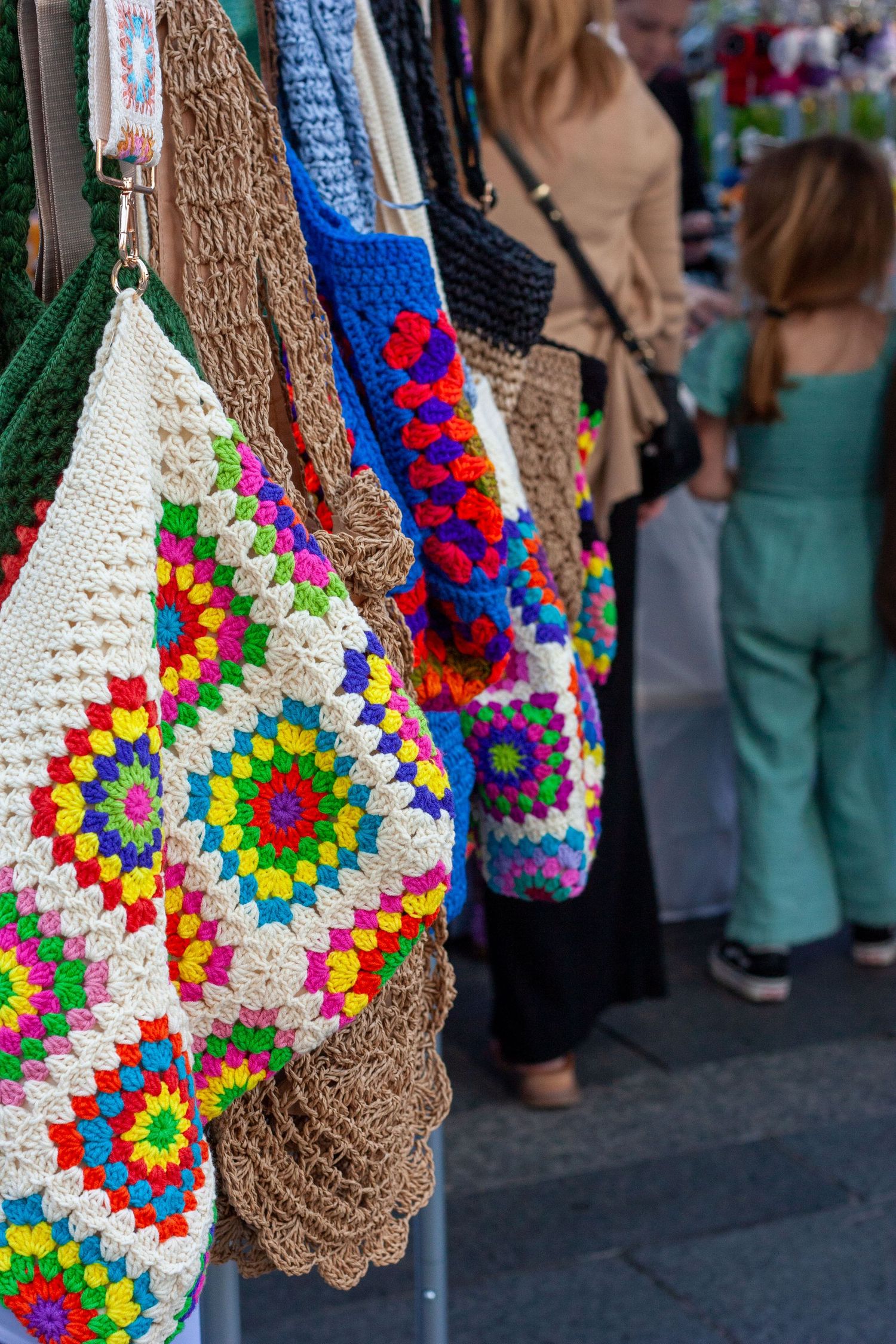 Colorful crochet granny square bags displayed at an outdoor market stall with shoppers browsing in the background.