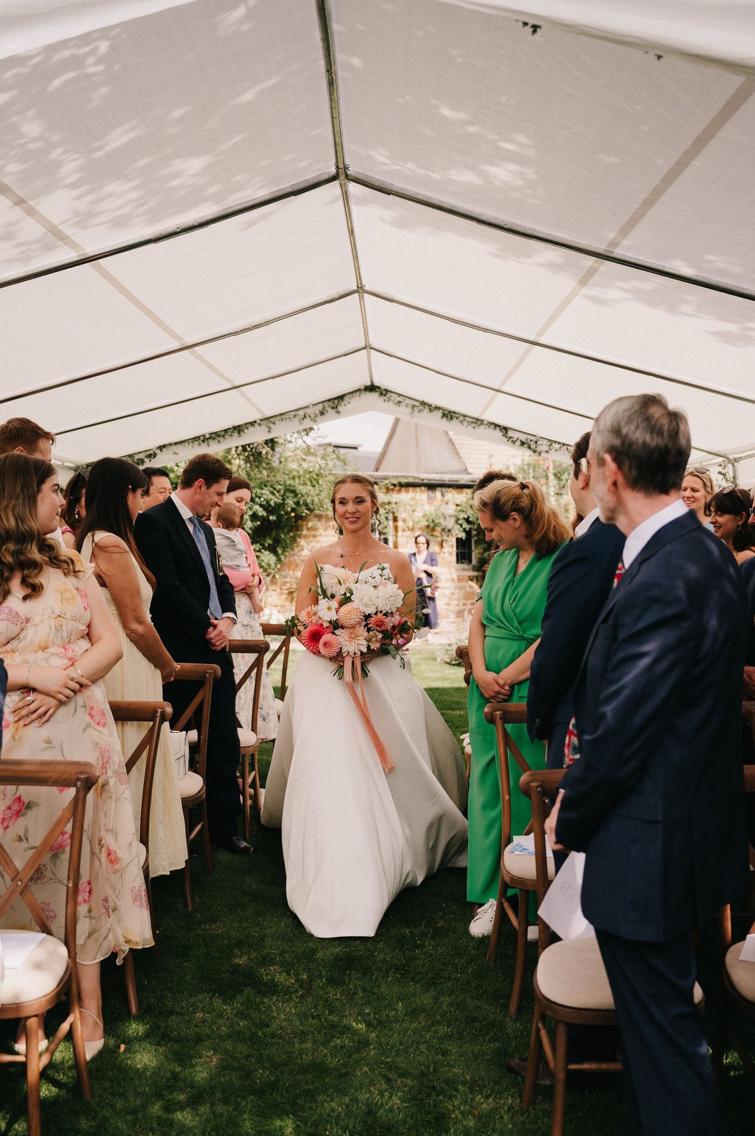 Smiling bride in white gown walks down the aisle under a marquee tent surrounded by seated wedding guests.