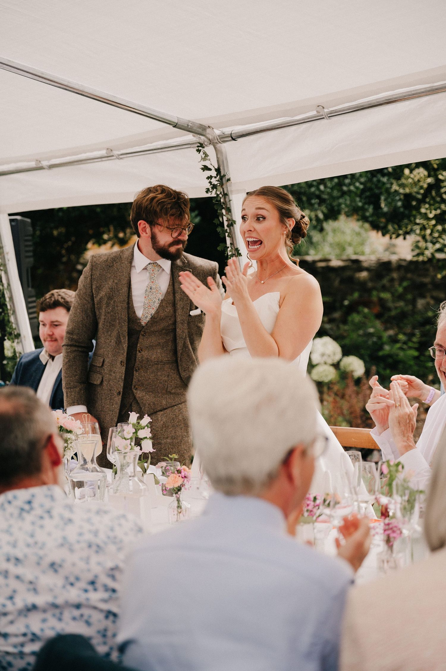 A bride and groom giving a speech under a marquee tent at their outdoor wedding reception with guests applauding.