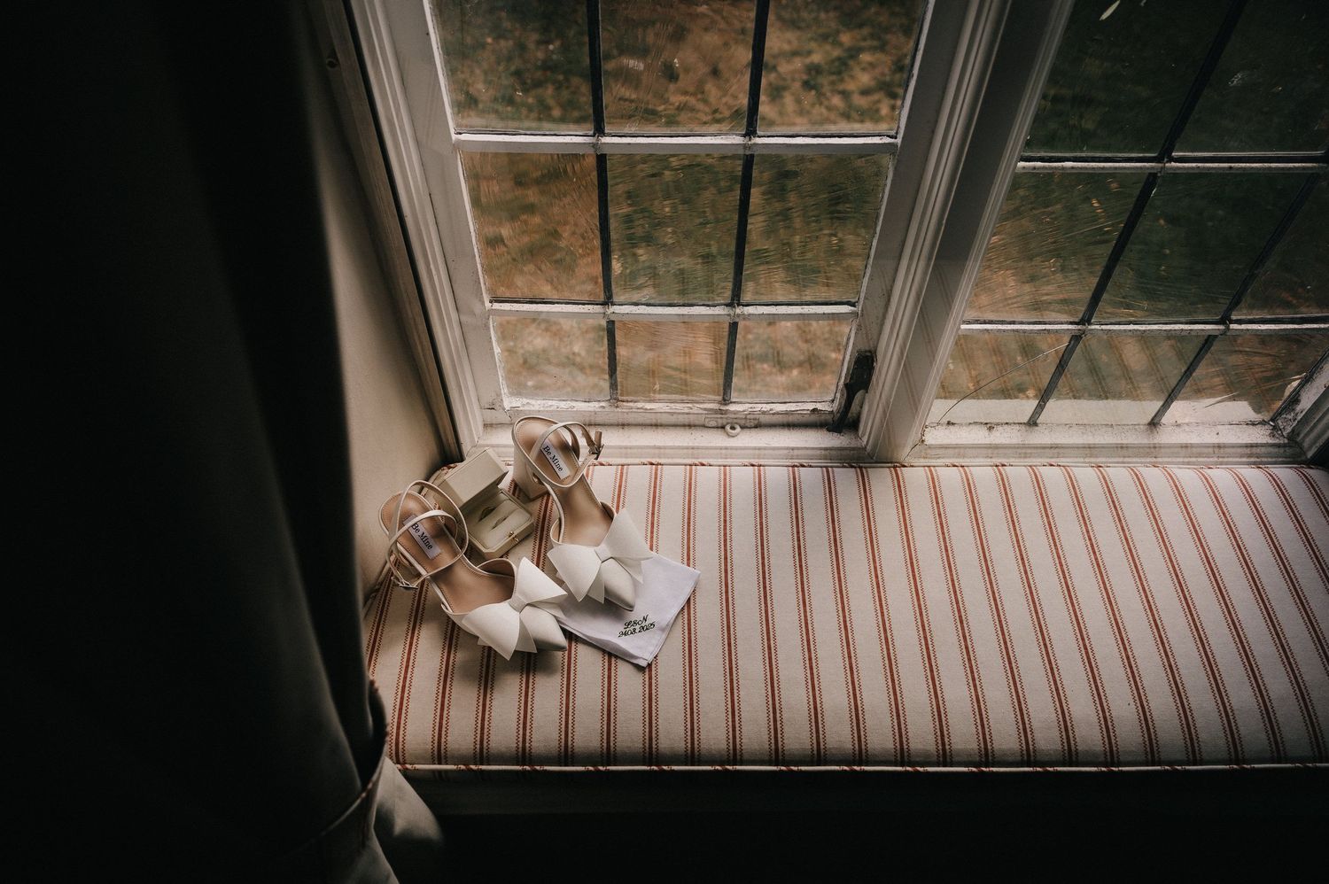 Elegant white bridal sandals resting on a striped window seat beside a rainy leaded glass window.