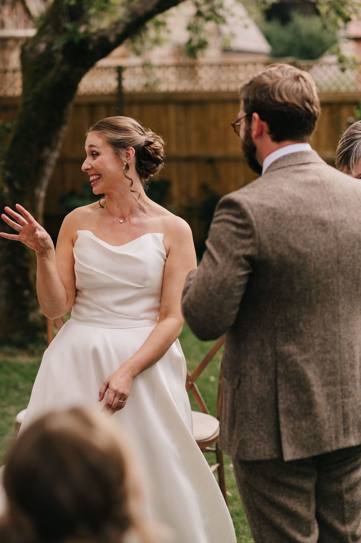 A smiling bride in a strapless white gown gestures during her outdoor wedding ceremony.