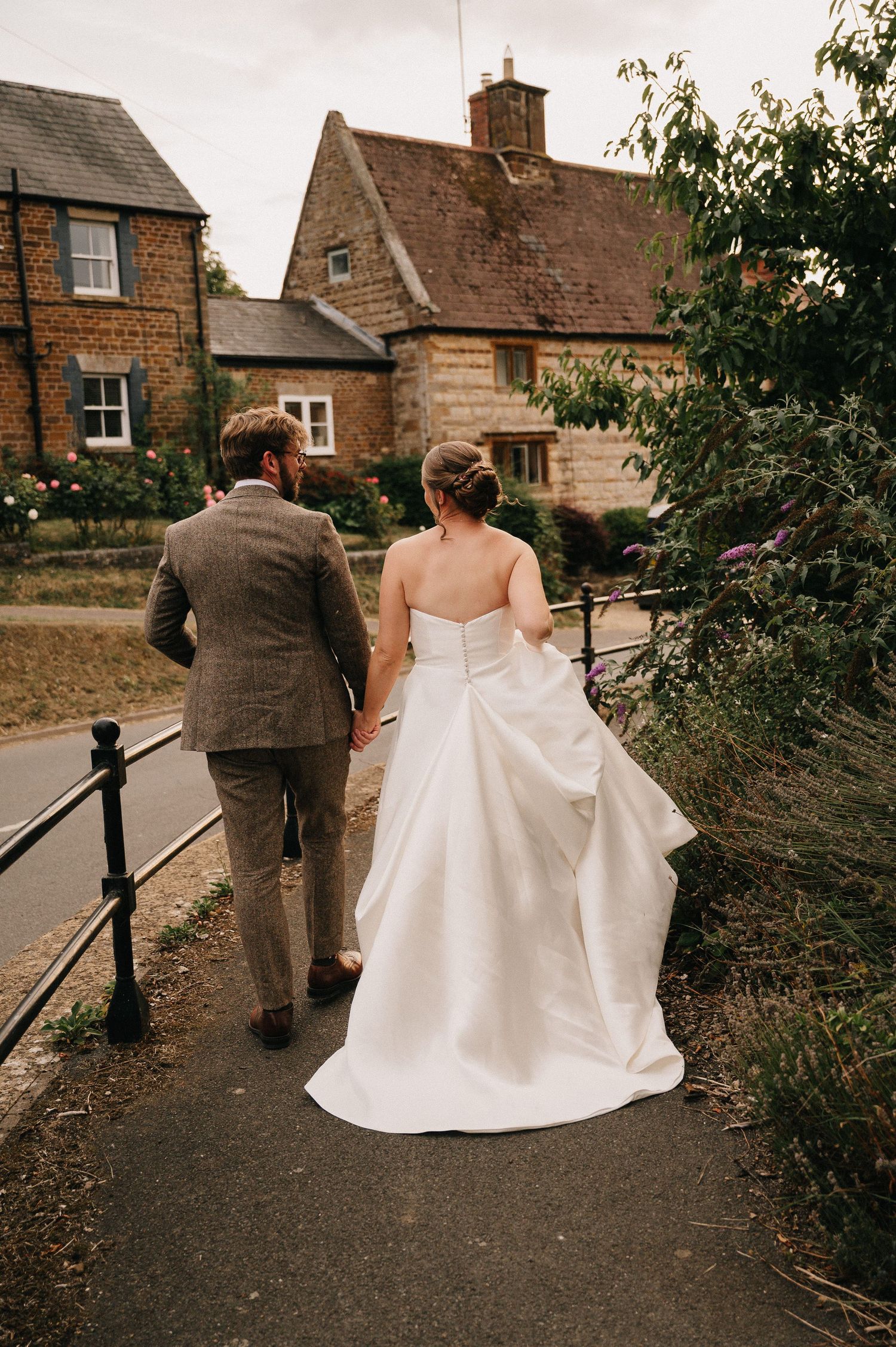 Bride and groom walking hand in hand down a charming village path, viewed from behind, in a white gown and tweed suit.