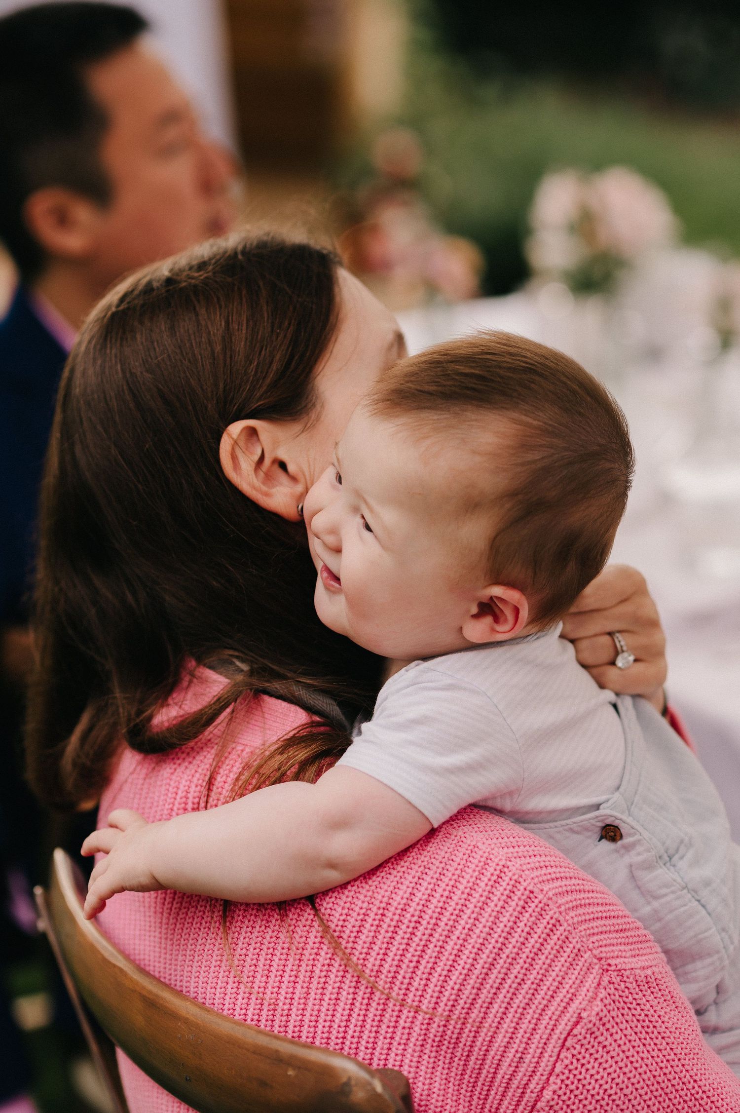 A mother tenderly embraces her smiling baby at an outdoor event, both sharing a sweet moment of joy.