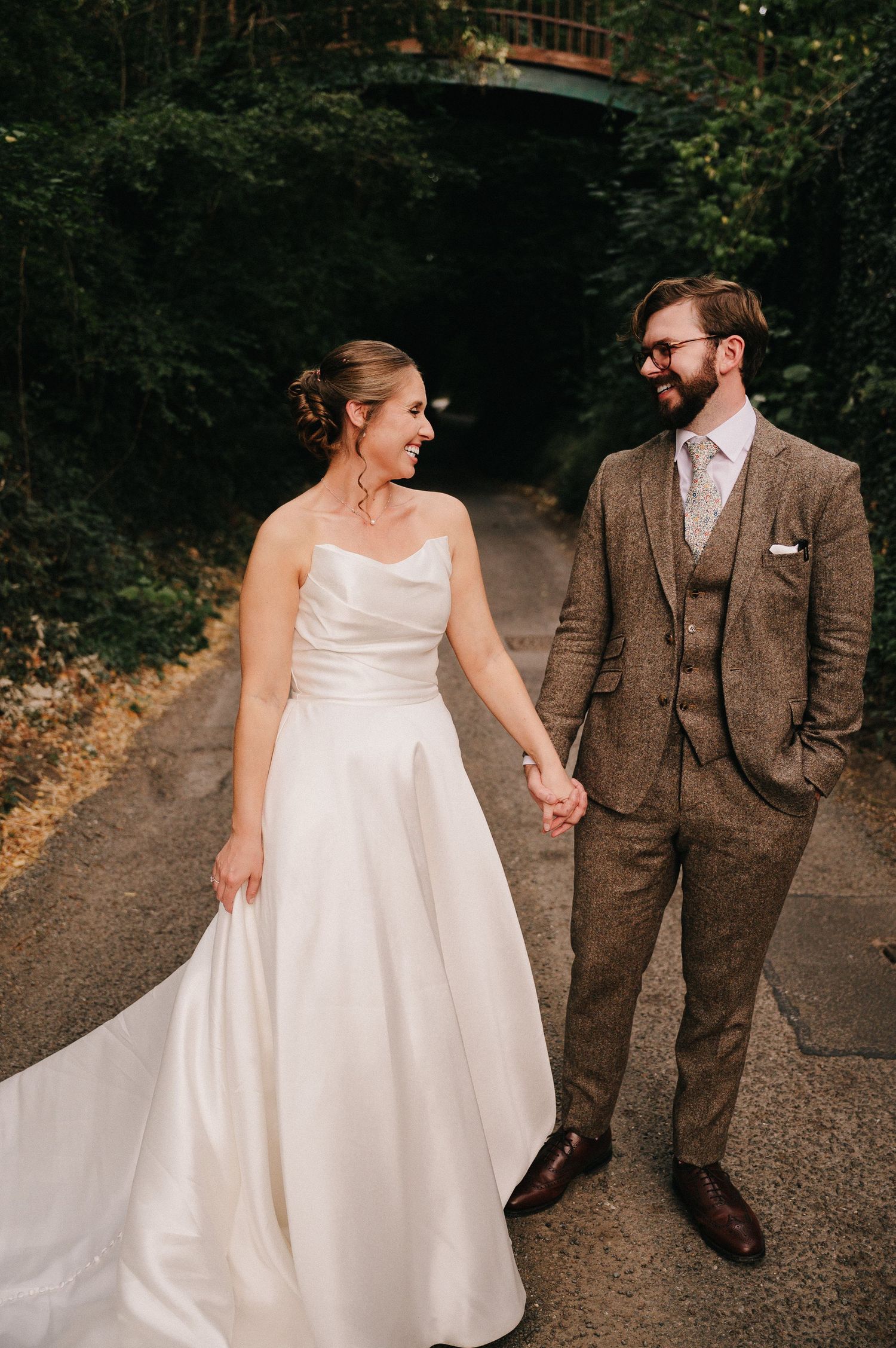 Bride and groom holding hands on a tree-lined path, she in a white gown, he in a brown tweed suit.