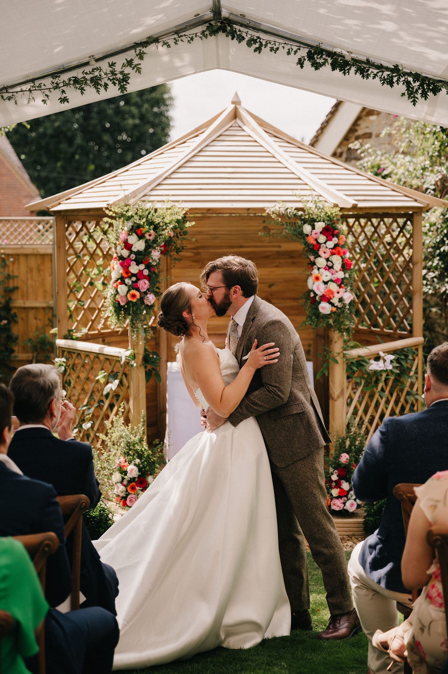 Bride and groom share first kiss at outdoor wedding ceremony beneath floral-adorned wooden gazebo with guests watching.