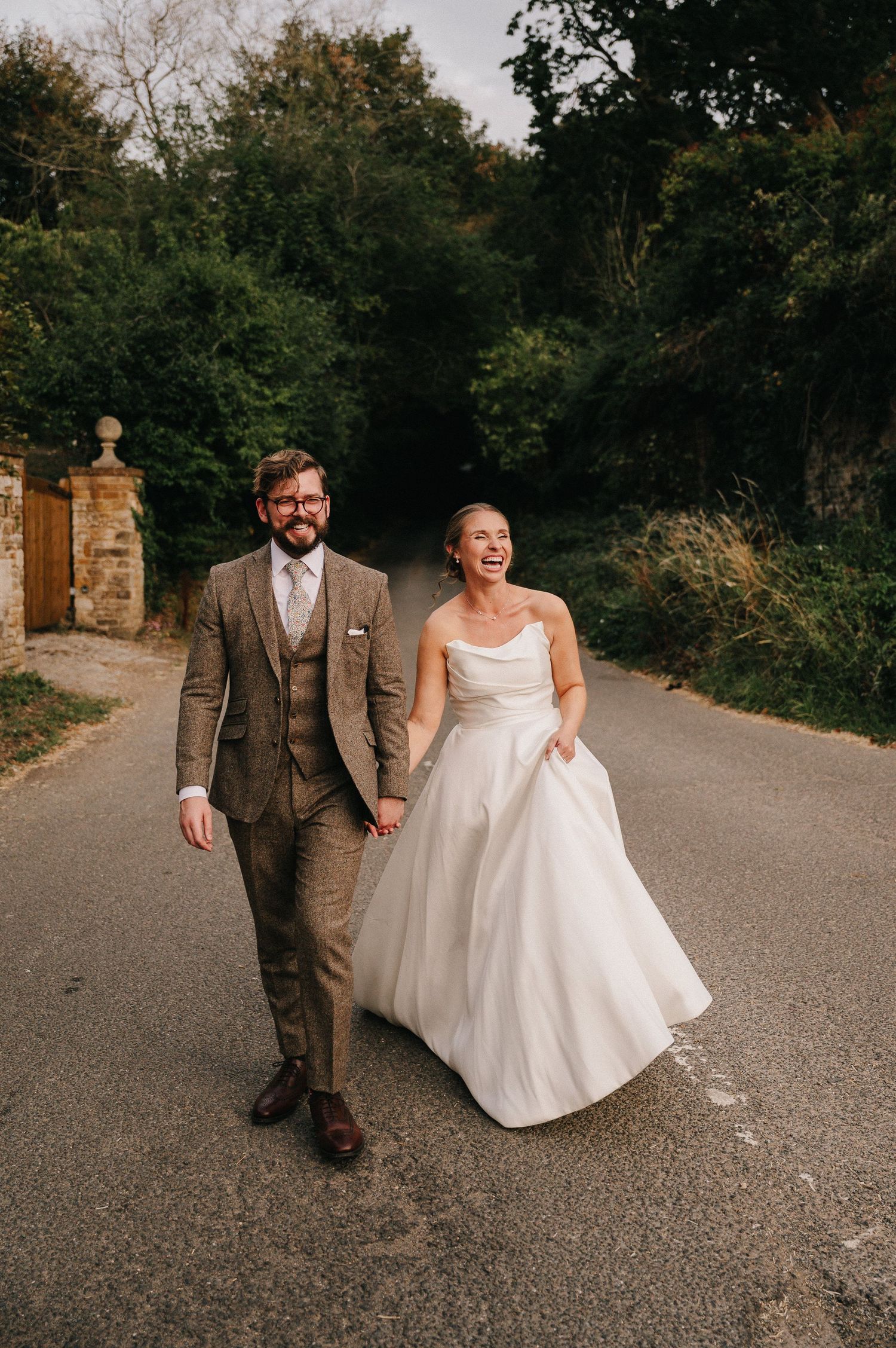 Joyful bride in white gown and groom in tweed suit walking hand in hand down a tree-lined road.