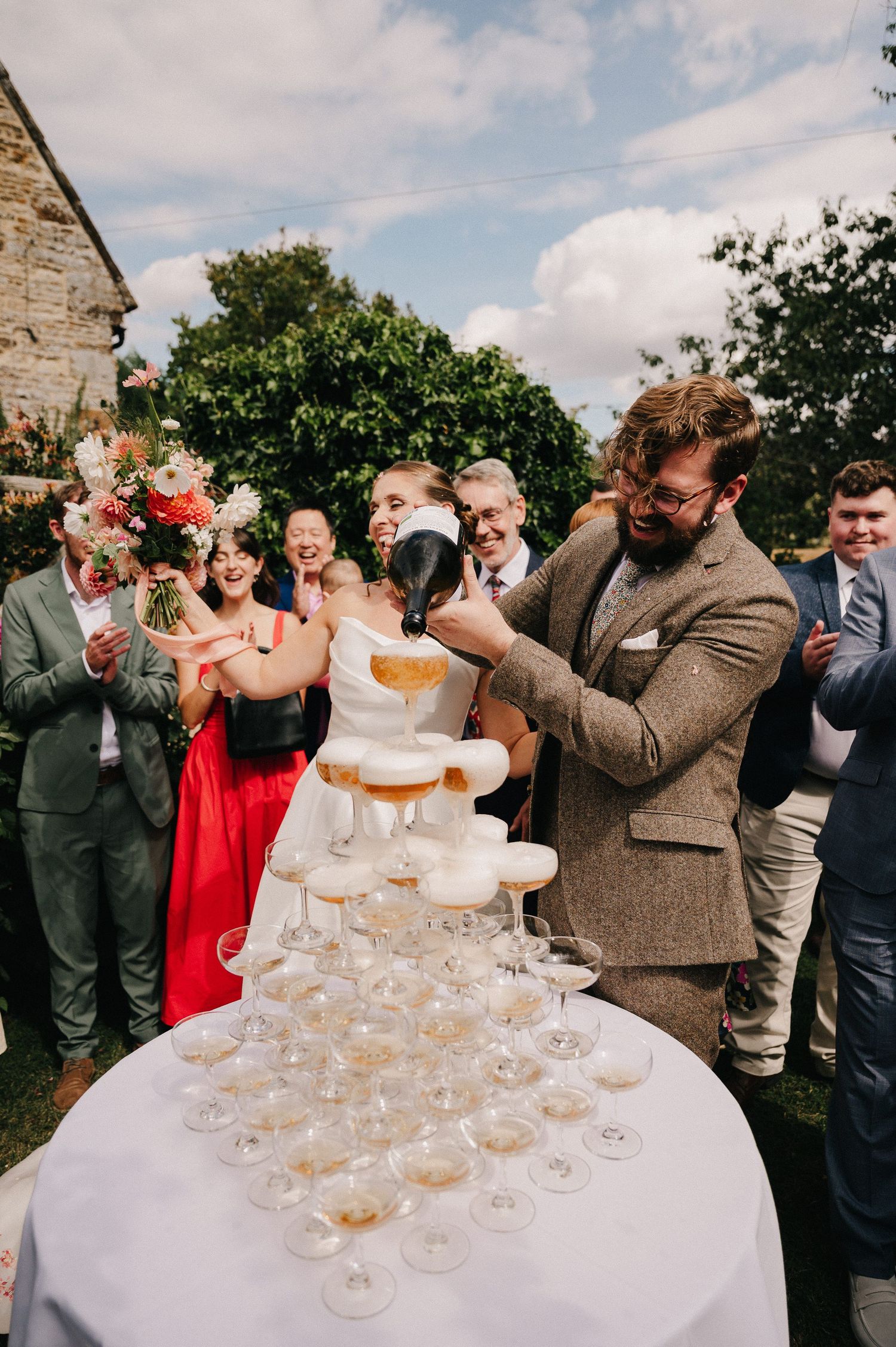 Bride and groom pouring champagne tower at outdoor wedding reception with guests celebrating in background.