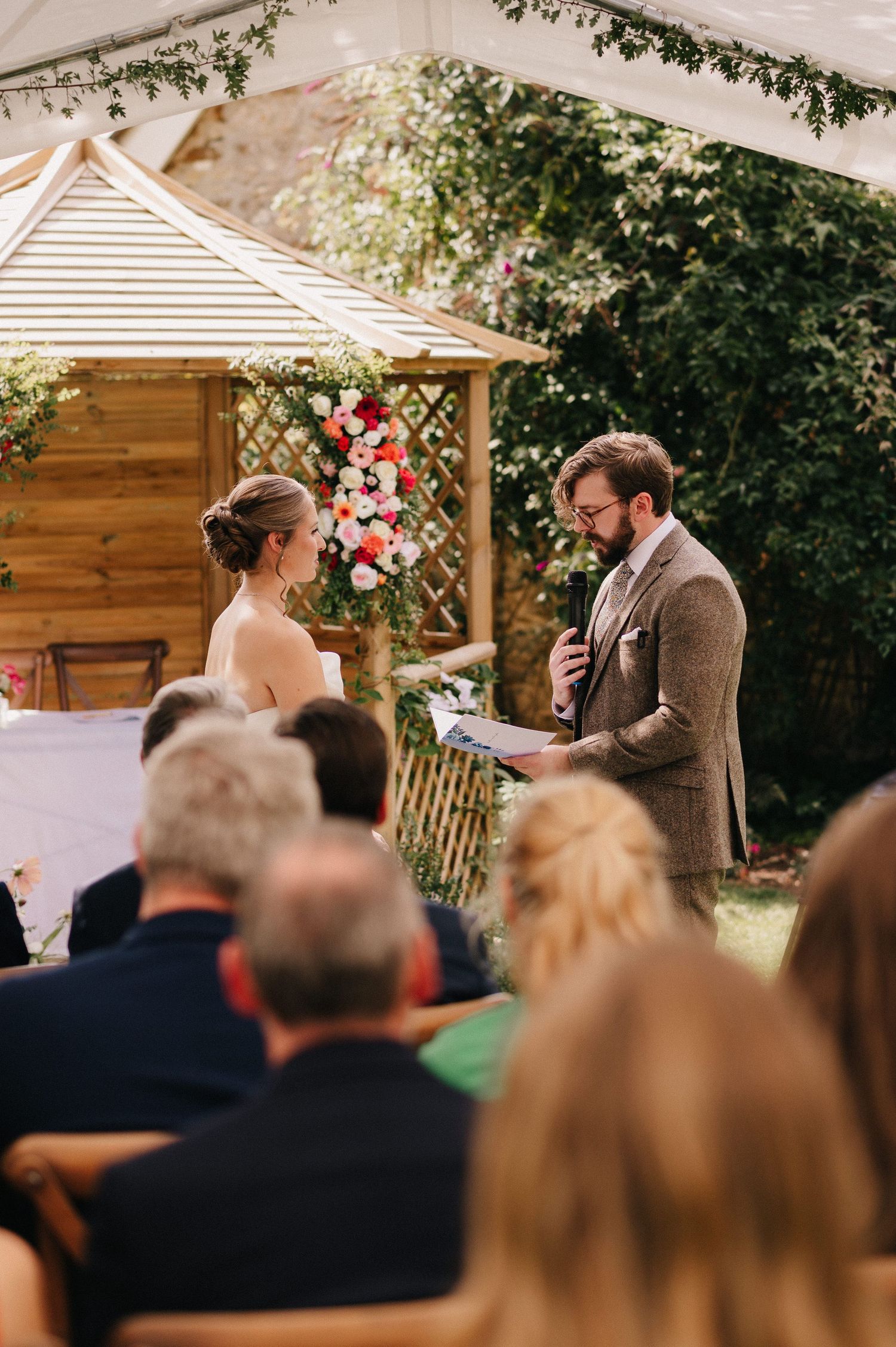 Groom reads vows to bride at outdoor wedding ceremony with floral arch backdrop and seated guests.