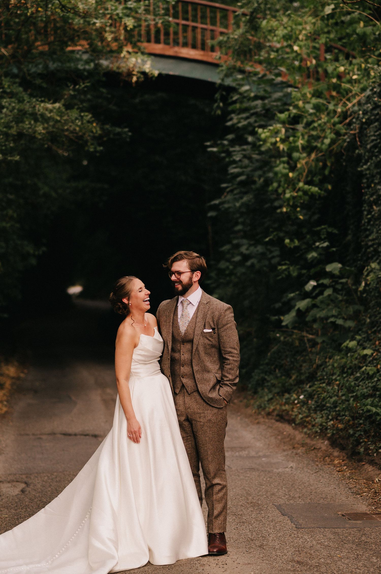 A bride in a white gown and groom in a brown suit stand on a tree-lined path beneath a bridge.