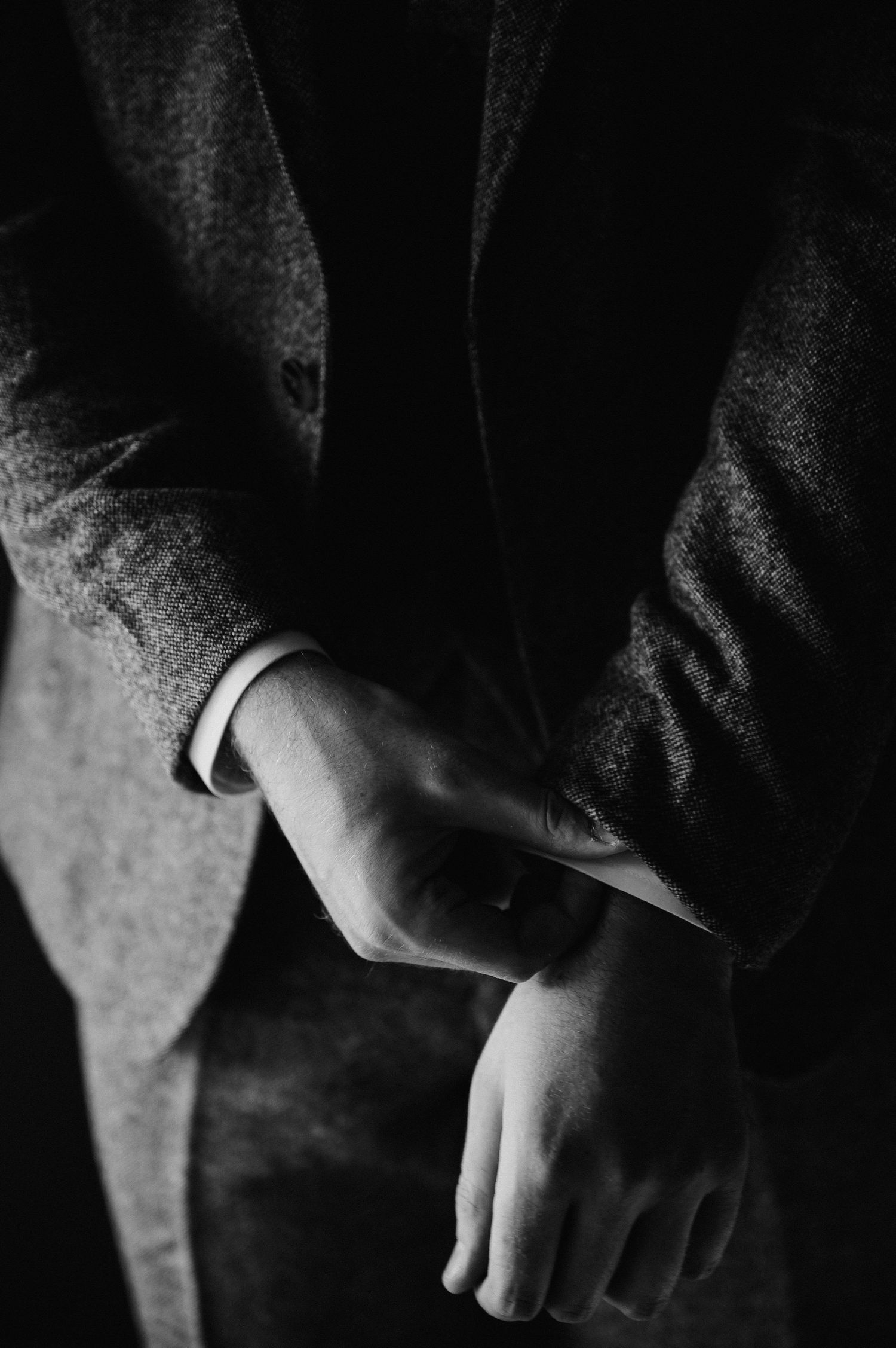 Black and white close-up of a man in a suit jacket adjusting his cufflinks, hands crossed at the wrist.
