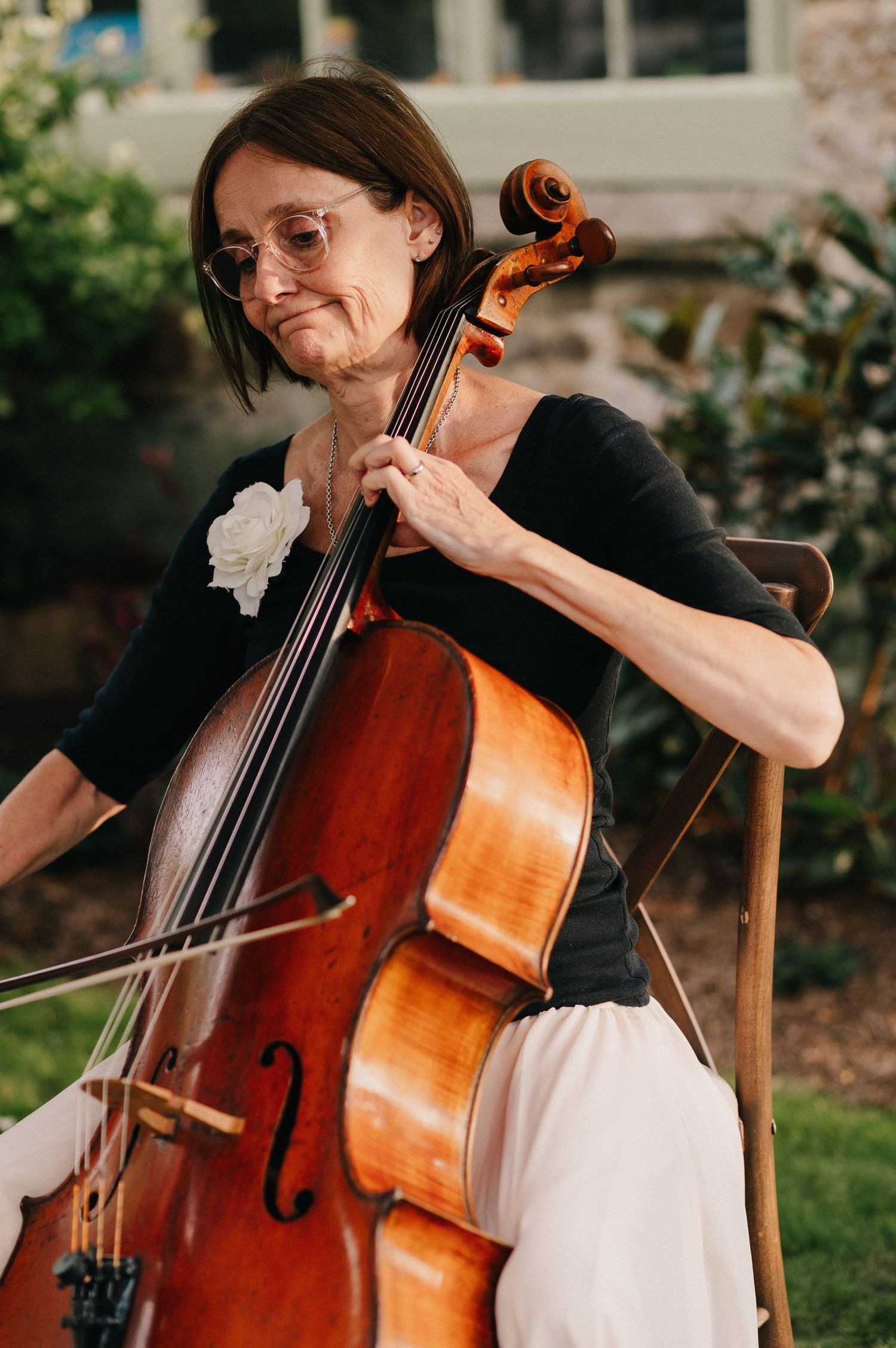 A woman playing cello outdoors, wearing glasses and a black top with a white flower, performing at a garden event.