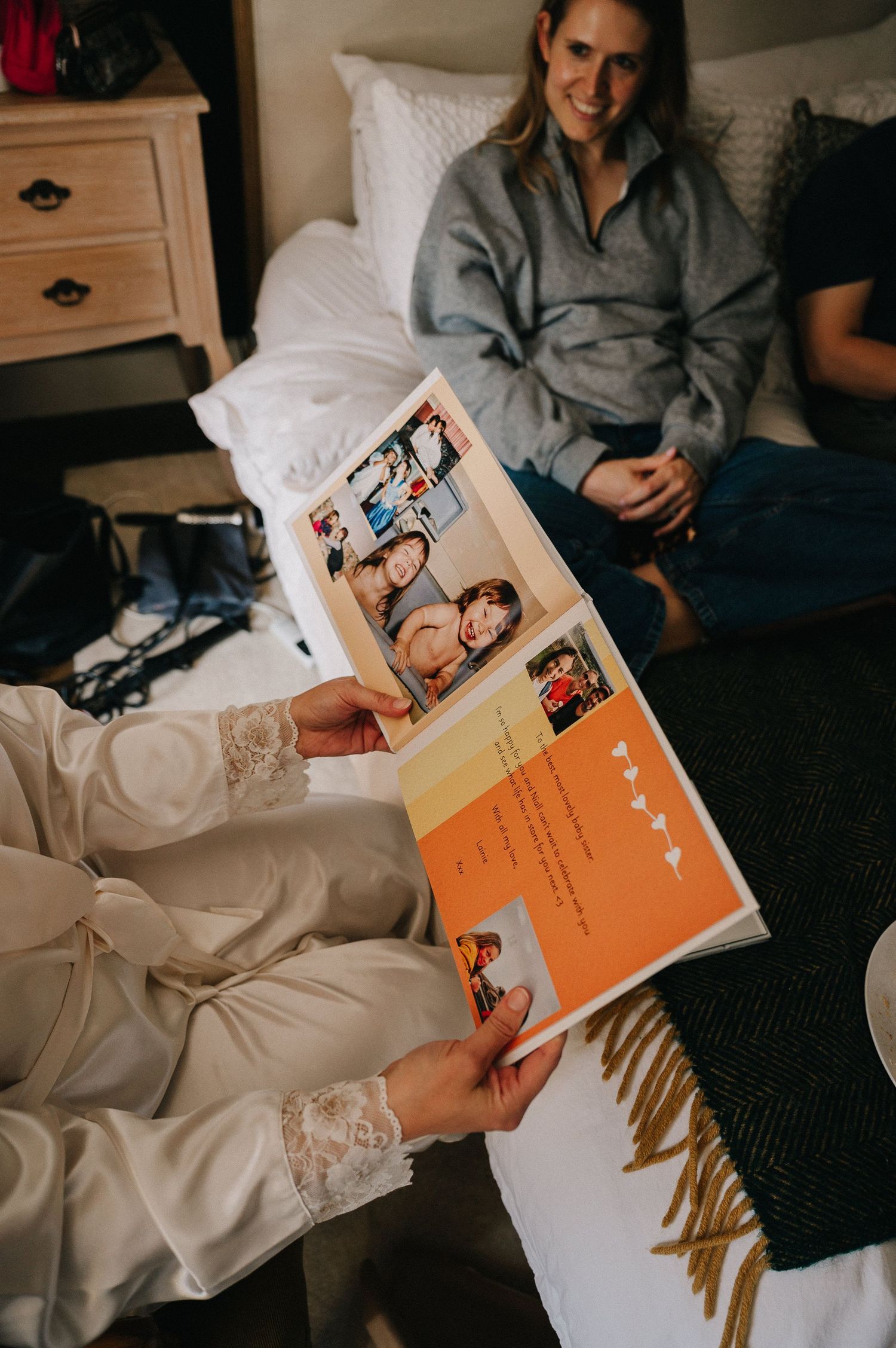Bride in white lace gown reads an orange photo book card while a smiling woman in gray sweater sits on bed nearby.