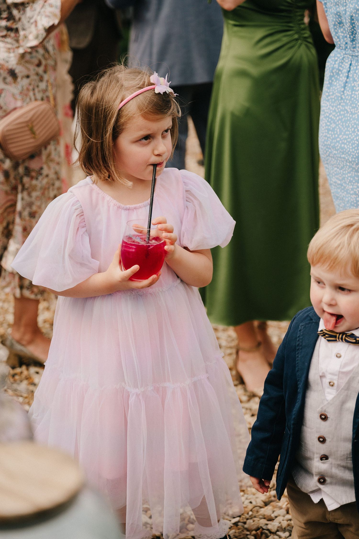 A young girl in a pink tulle dress sips a red drink through a straw at a wedding celebration outdoors.