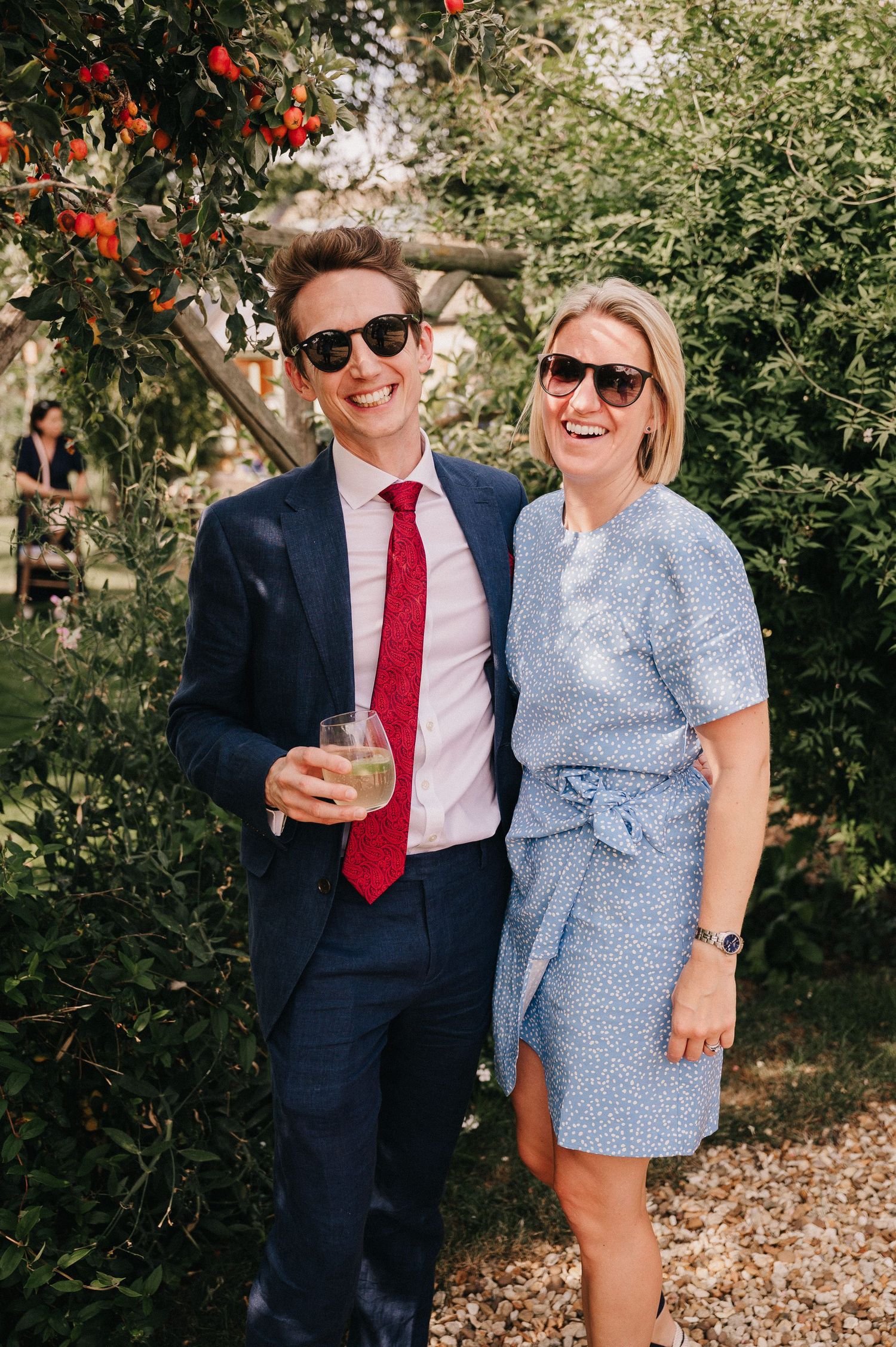 Smiling couple in sunglasses at outdoor garden party, man in navy suit with red tie, woman in blue floral dress.