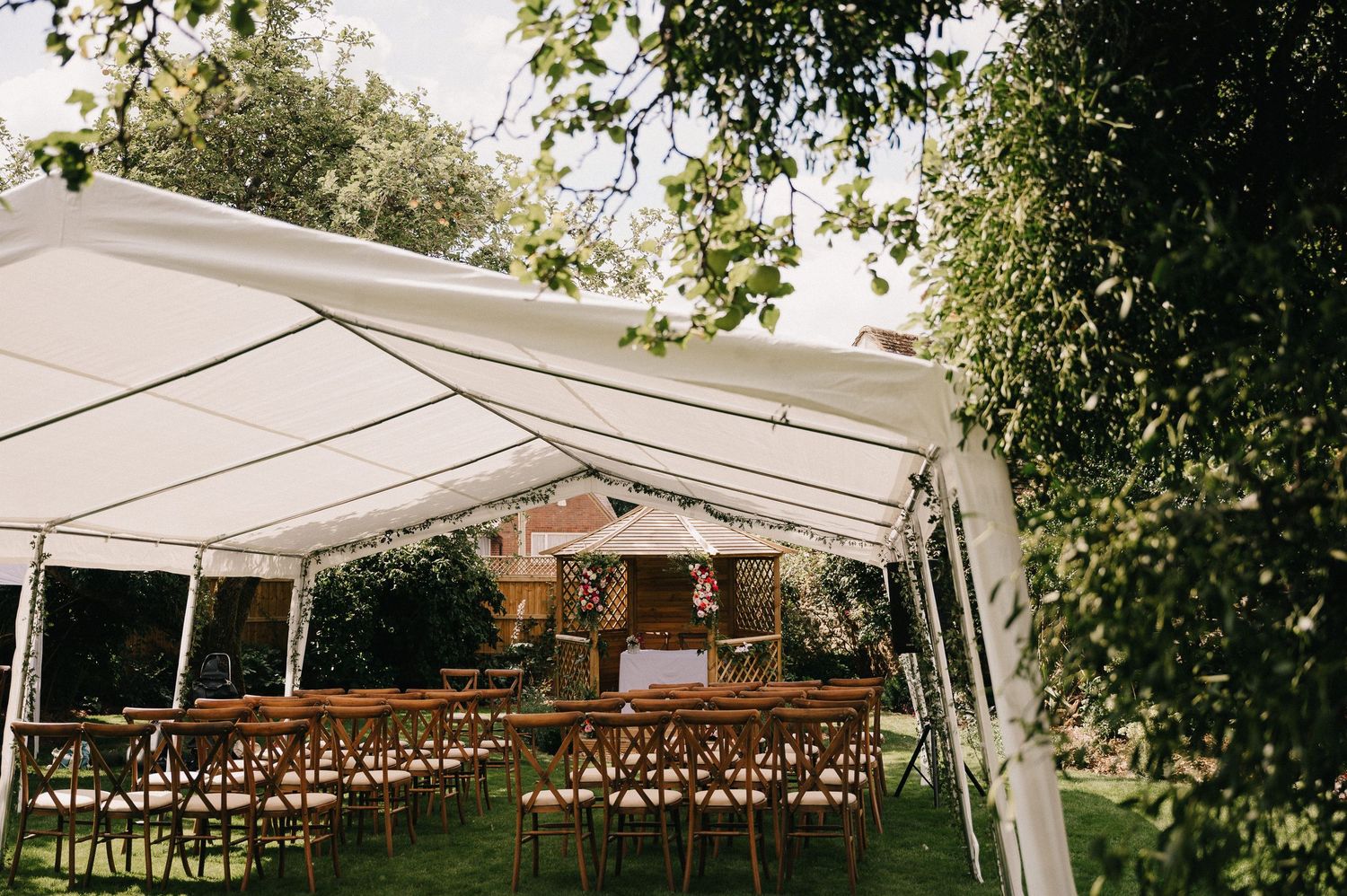 Elegant outdoor wedding ceremony setup under a white marquee tent with wooden cross-back chairs on lush green lawn.