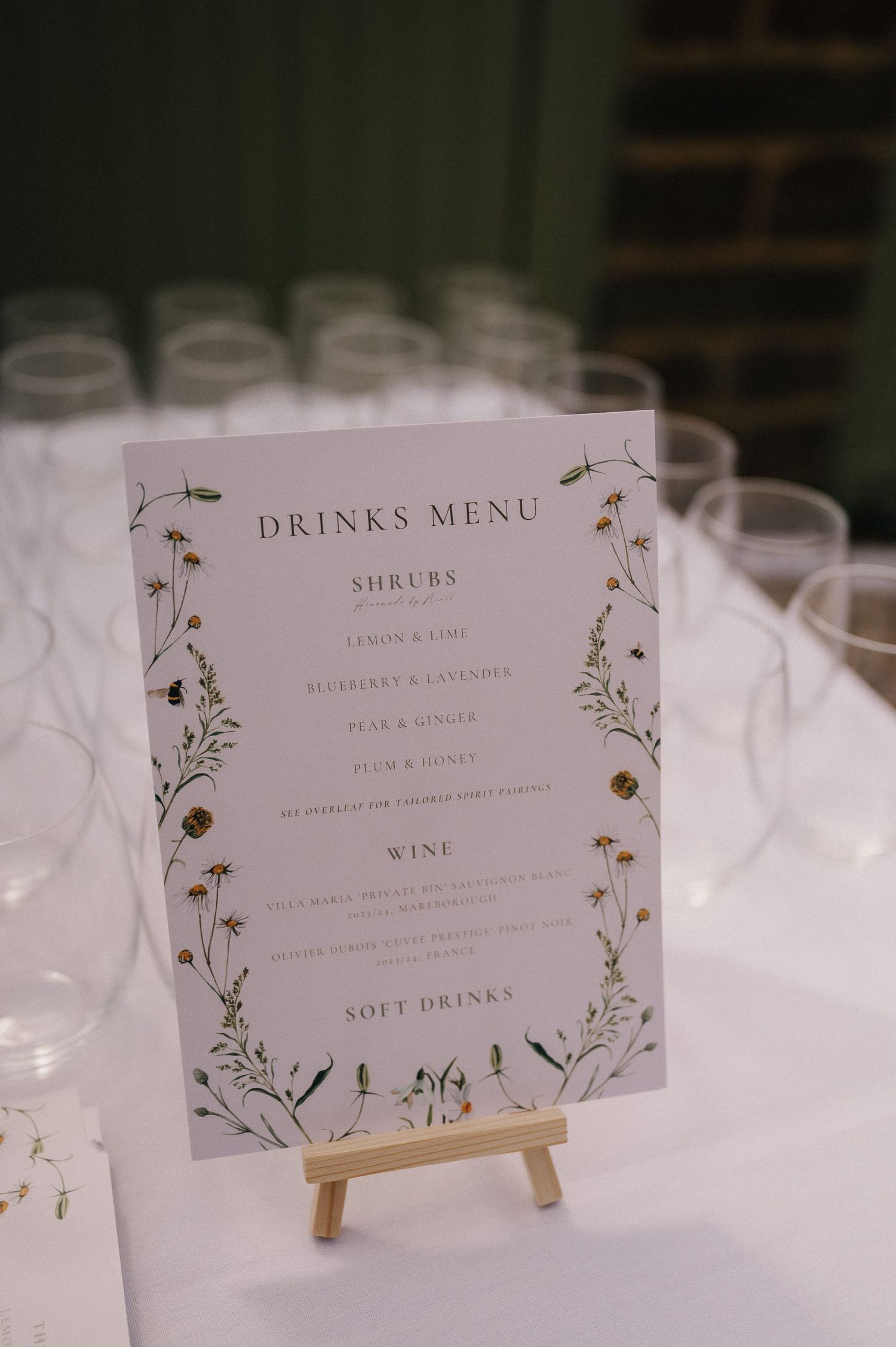 Elegant wedding drinks menu on easel featuring shrubs, wine, and soft drinks with floral botanical border design.