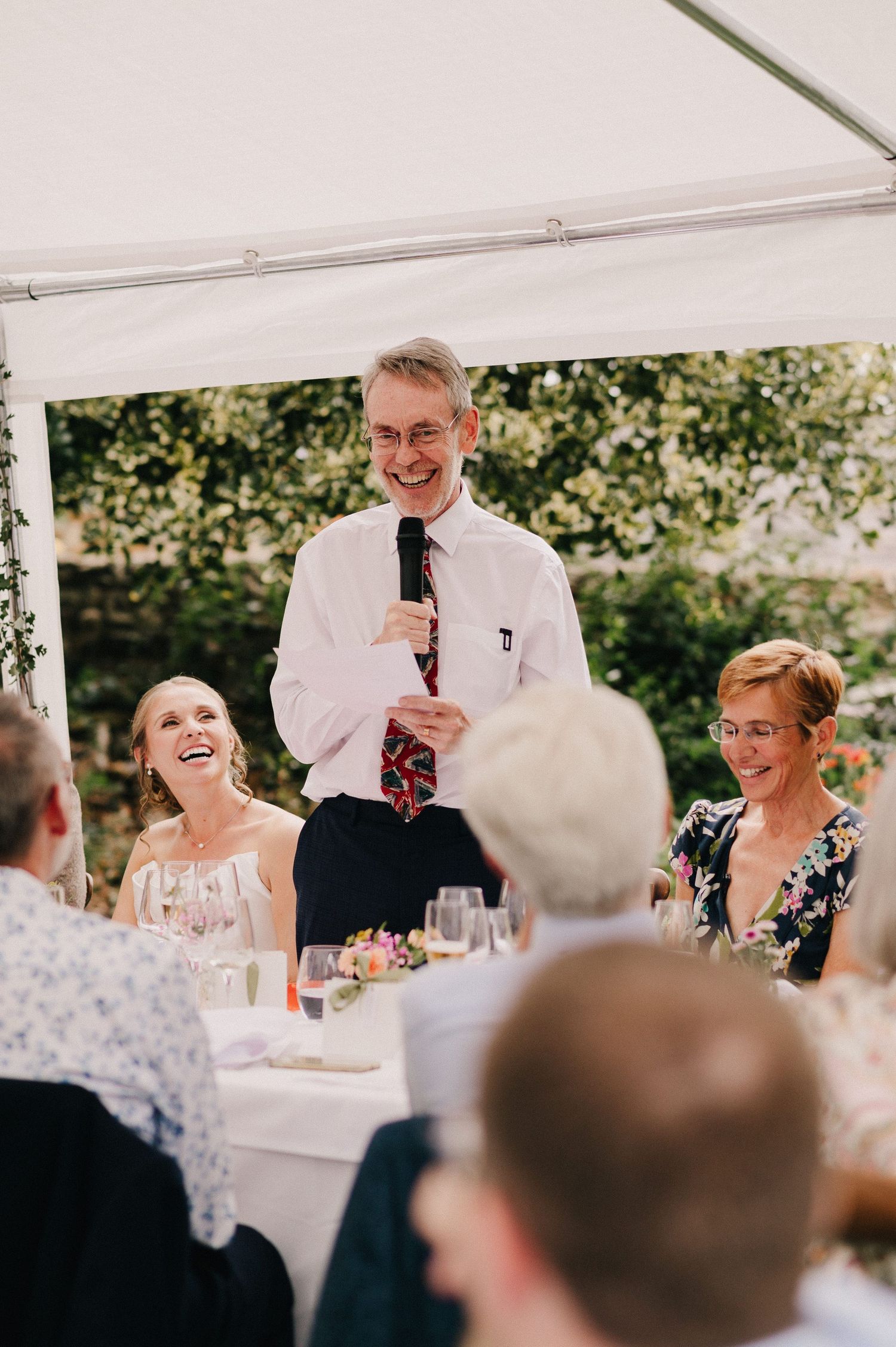 Man giving a speech at an outdoor wedding reception under a white tent, guests laughing and smiling around a decorated table.