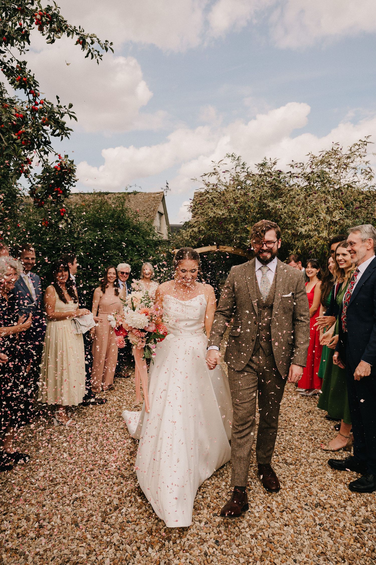 Bride and groom walk through confetti shower surrounded by guests outdoors at rustic wedding venue.