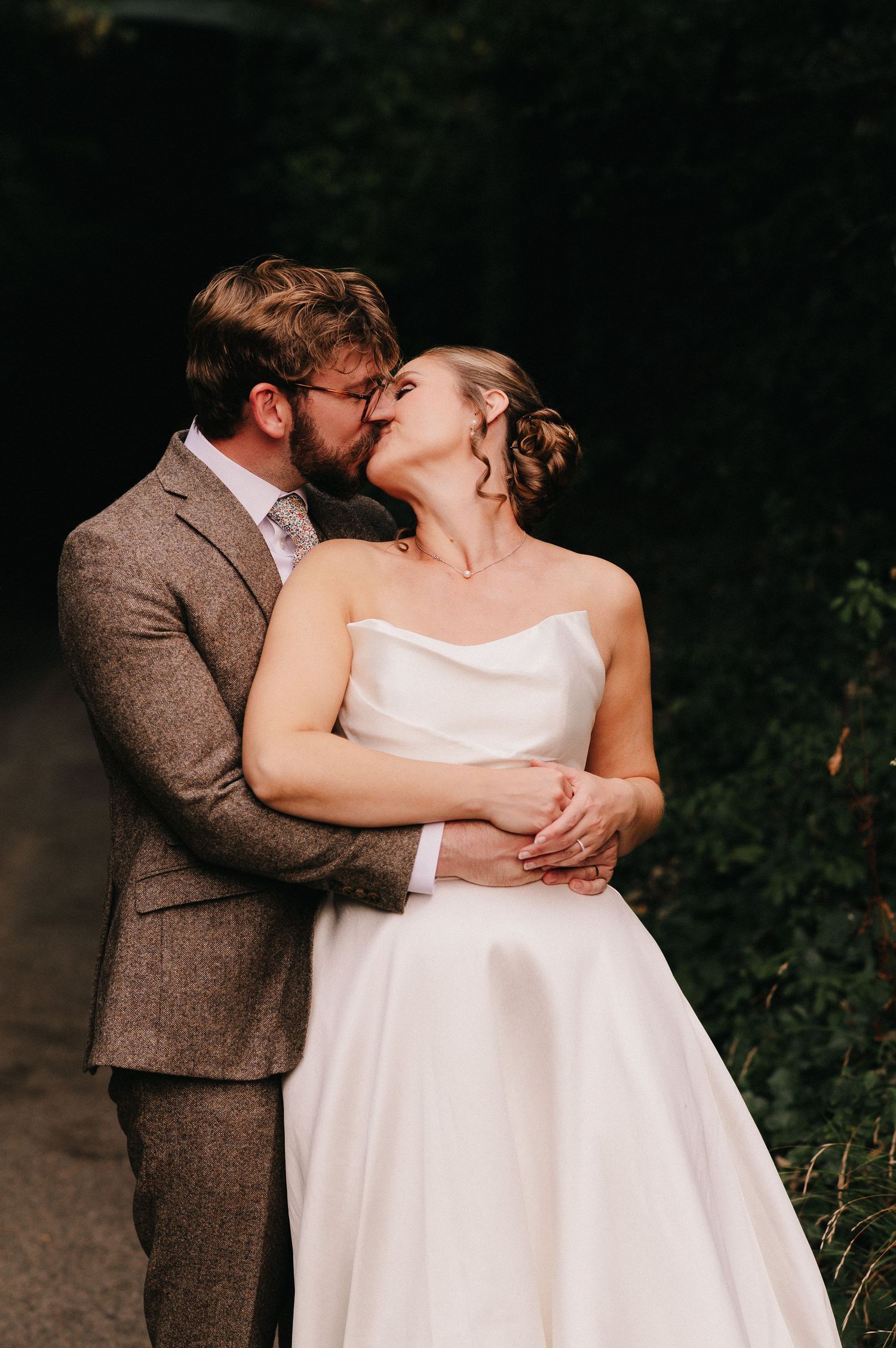 A groom in a tweed suit kisses his bride in a strapless white gown outdoors amid lush greenery.