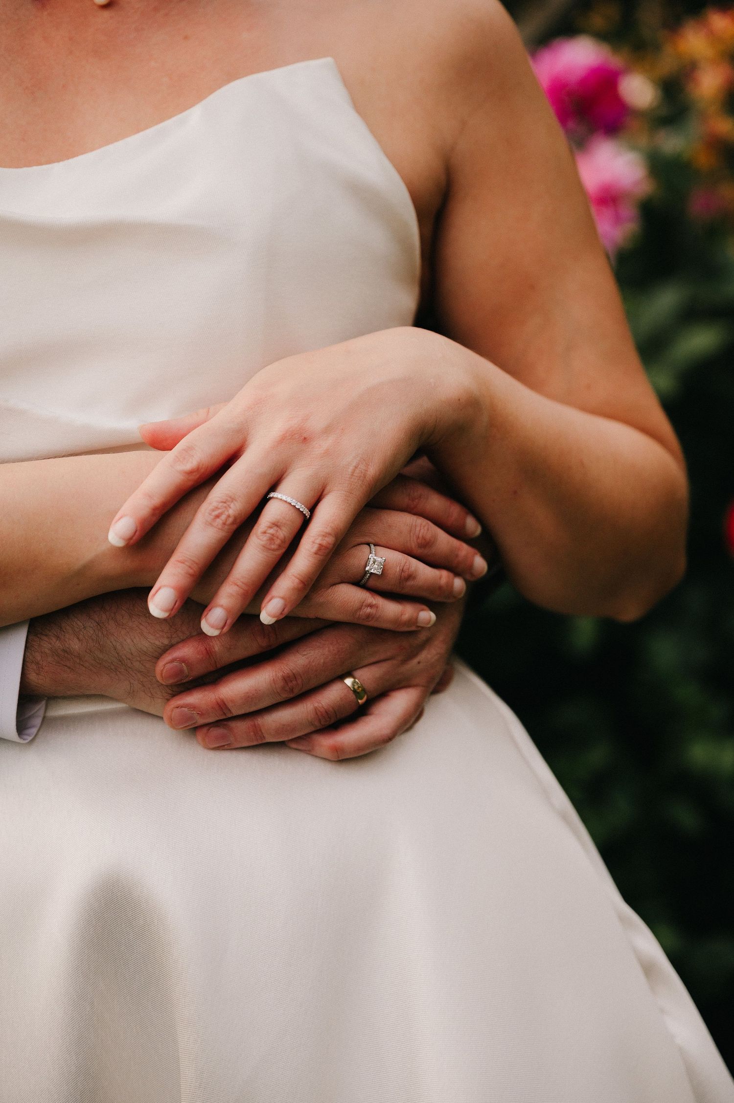 Close-up of newlyweds hands showing wedding rings, bride in white dress embraced by groom.