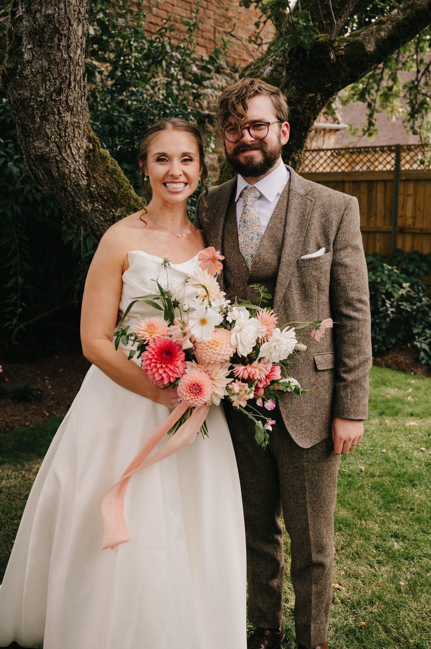 Smiling bride in white strapless gown holds coral bouquet beside groom in brown tweed suit outdoors.