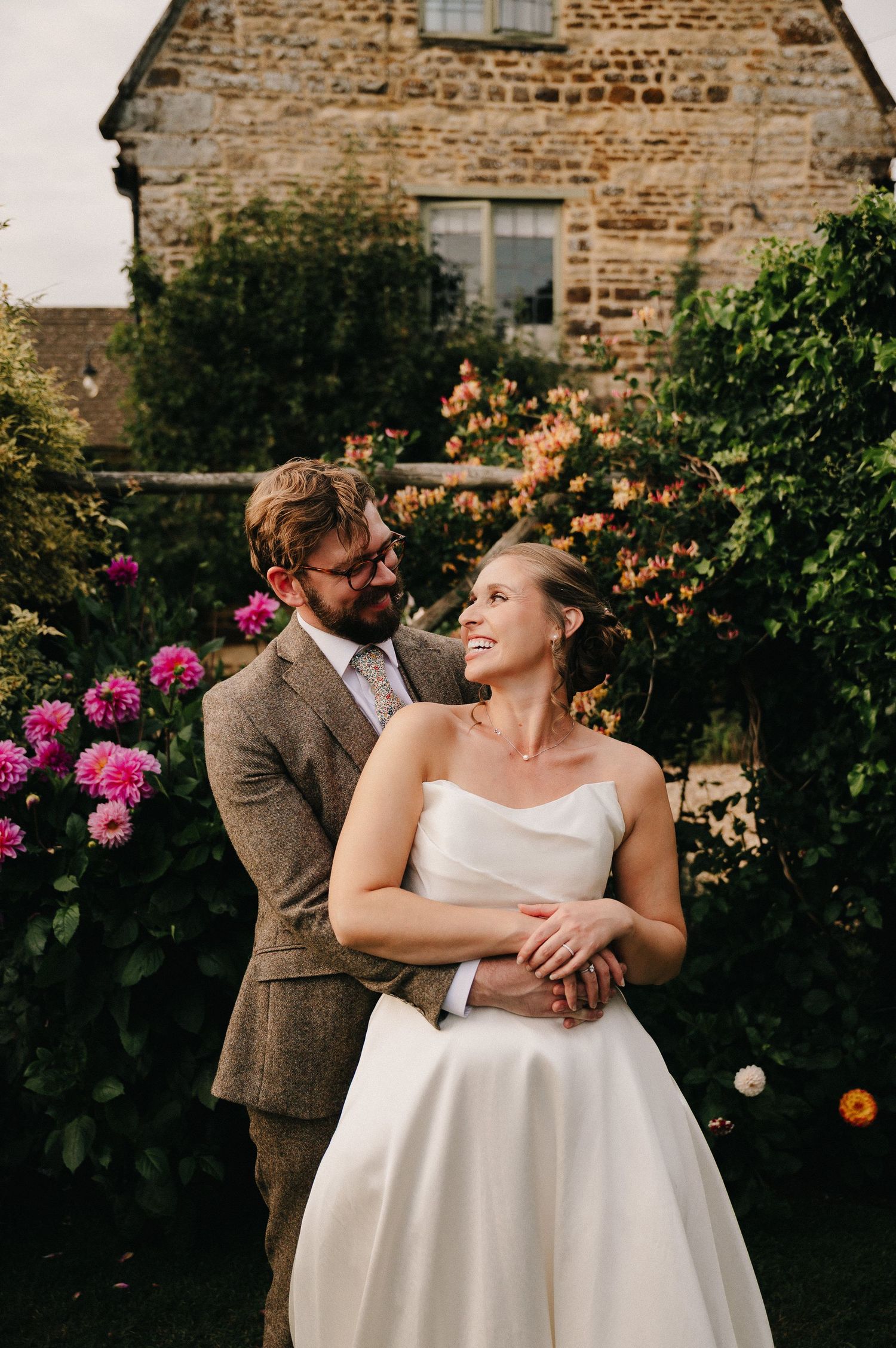 Bride and groom embrace in a lush garden beside a stone building, surrounded by pink and orange blooms.