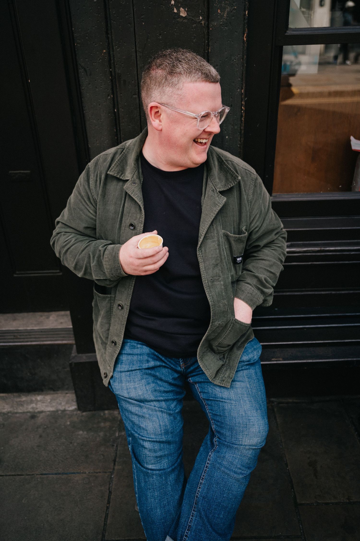 Man in green jacket and jeans laughing while holding a coffee cup outdoors.