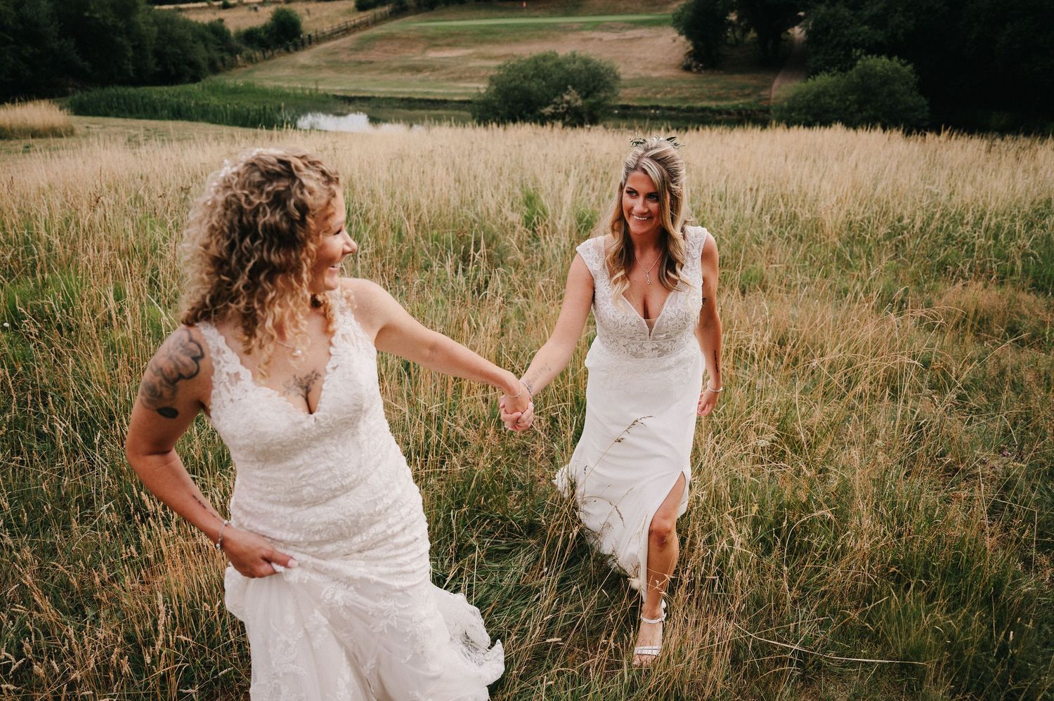 Two brides in white lace wedding dresses hold hands and walk through a lush green meadow on their wedding day.